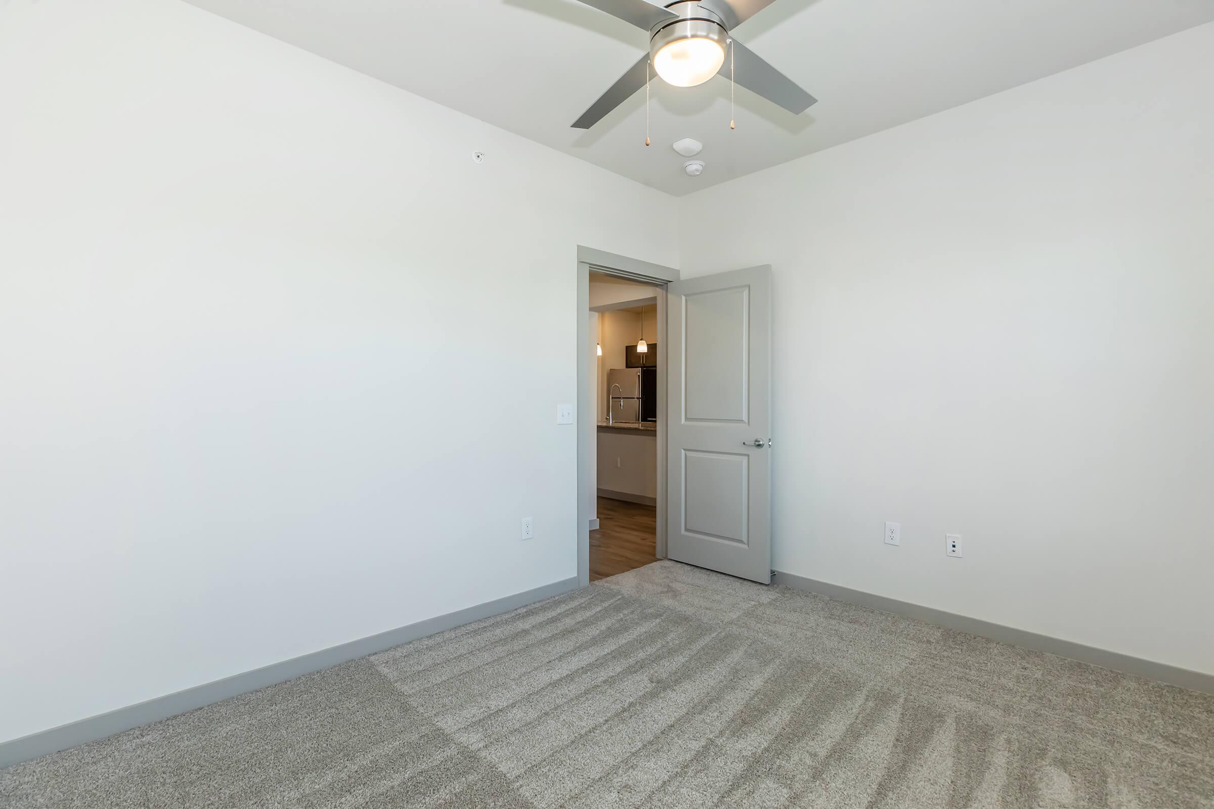 A spacious, empty room with light-colored walls and carpeted flooring. A ceiling fan is visible, and a doorway leads to a kitchen area in the background. The room is well-lit, giving it an open and inviting feel.