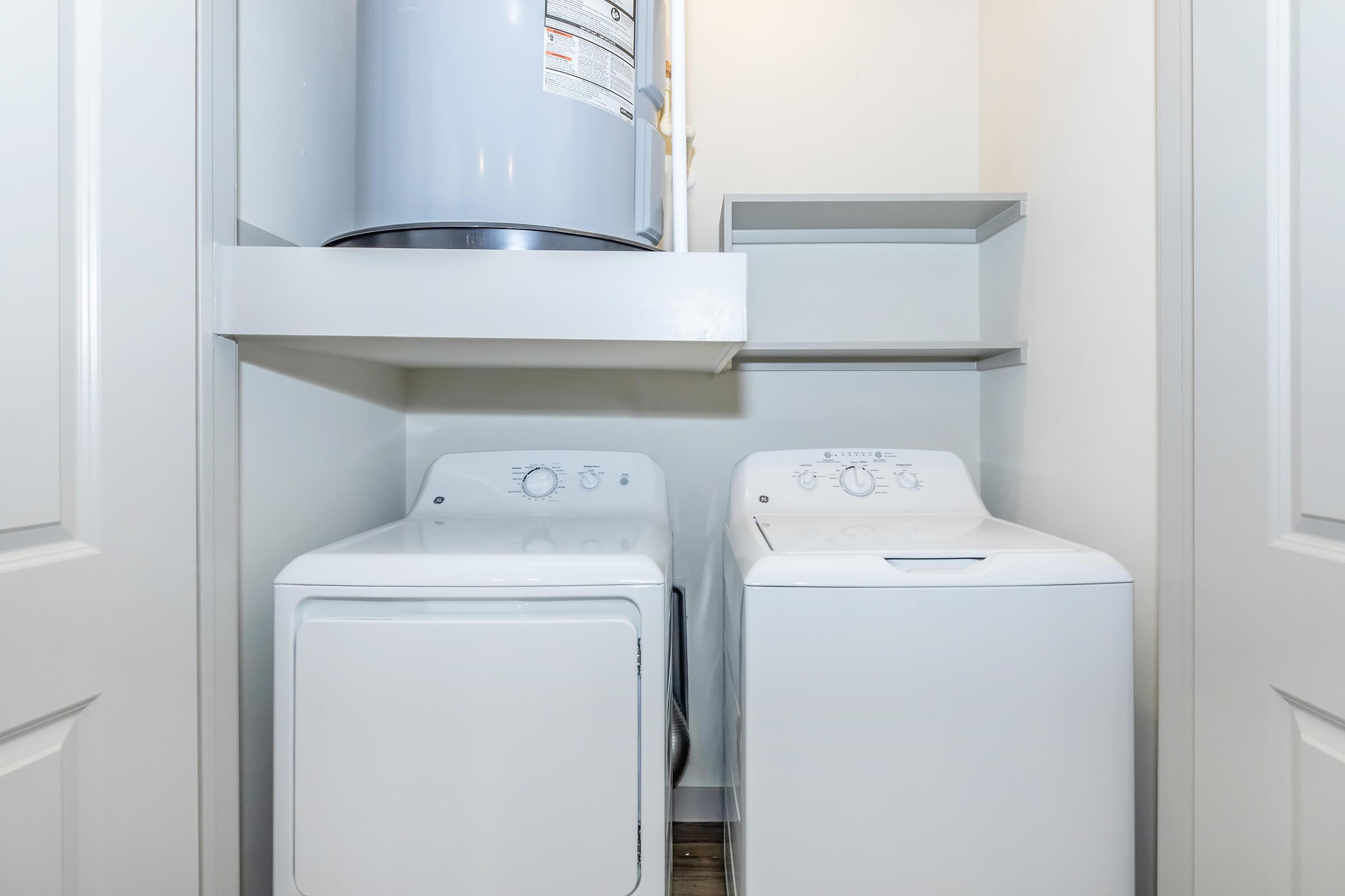 A compact laundry area featuring a stacked washing machine and dryer side by side. Above them, there is a shelf for storage. The walls are painted in a light color, and the floor is covered with wood-like tiles, creating a clean and functional space for laundry activities.