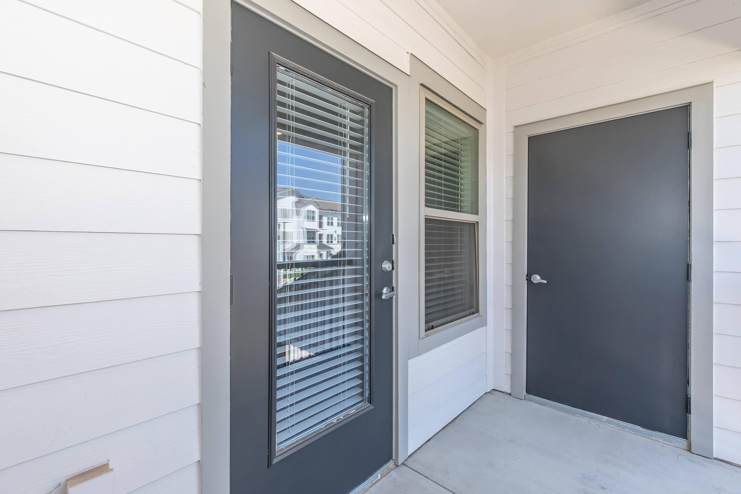 A modern entryway featuring two doors with contemporary gray frames. One door has vertical blinds and a large window beside it, while the other door is solid gray. The walls are white with a clean, minimalist aesthetic, and sunlight illuminates the area from an outside view.
