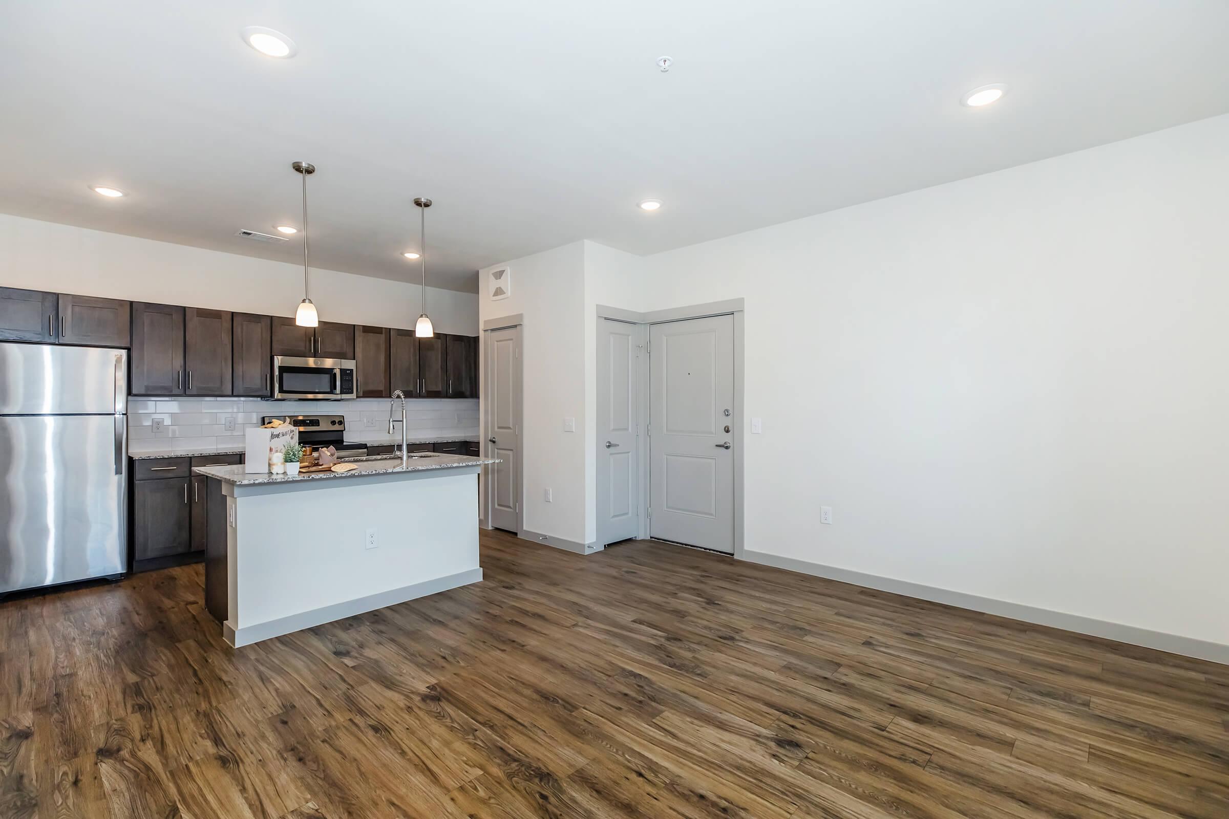 A modern kitchen and living area featuring stainless steel appliances, dark wooden cabinetry, and hardwood-style floors. The space includes a countertop with bar seating and an entrance door leading into the room. Bright overhead lights illuminate the area, creating a welcoming atmosphere.