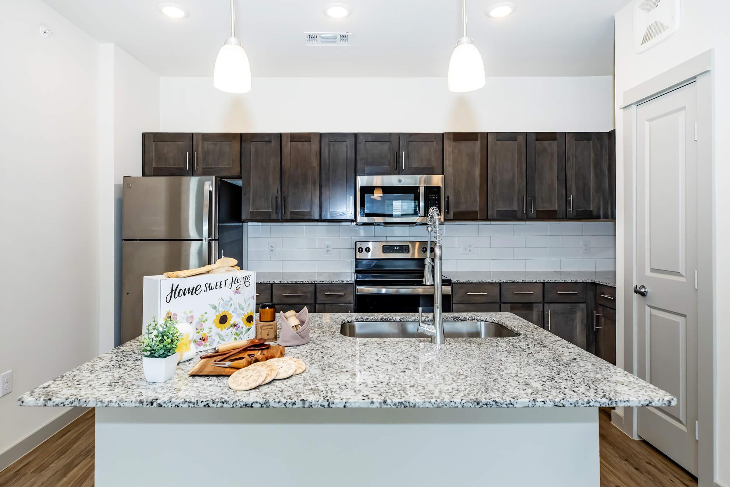 Modern kitchen featuring dark wood cabinetry, stainless steel appliances, and a large granite island. The island is decorated with baking items and a decorative sign. Bright pendant lights hang above, and the space is well-lit with natural light.