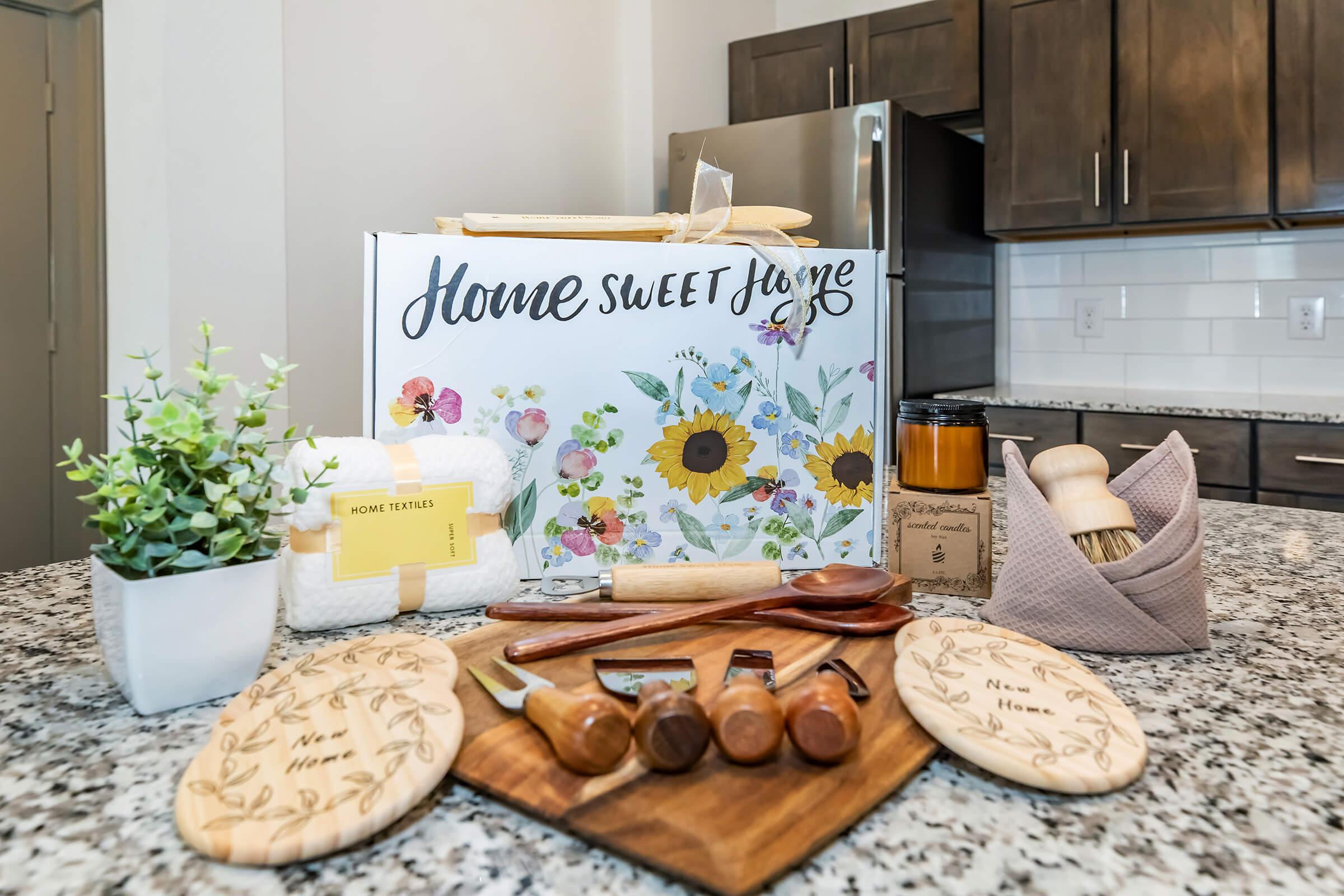 A floral-themed box labeled "Home Sweet Home" sits on a granite countertop. Surrounding it are various kitchen items, including engraved wooden boards, cooking utensils, a jar candle, a plant, and a dish towel, creating a cozy home ambiance.