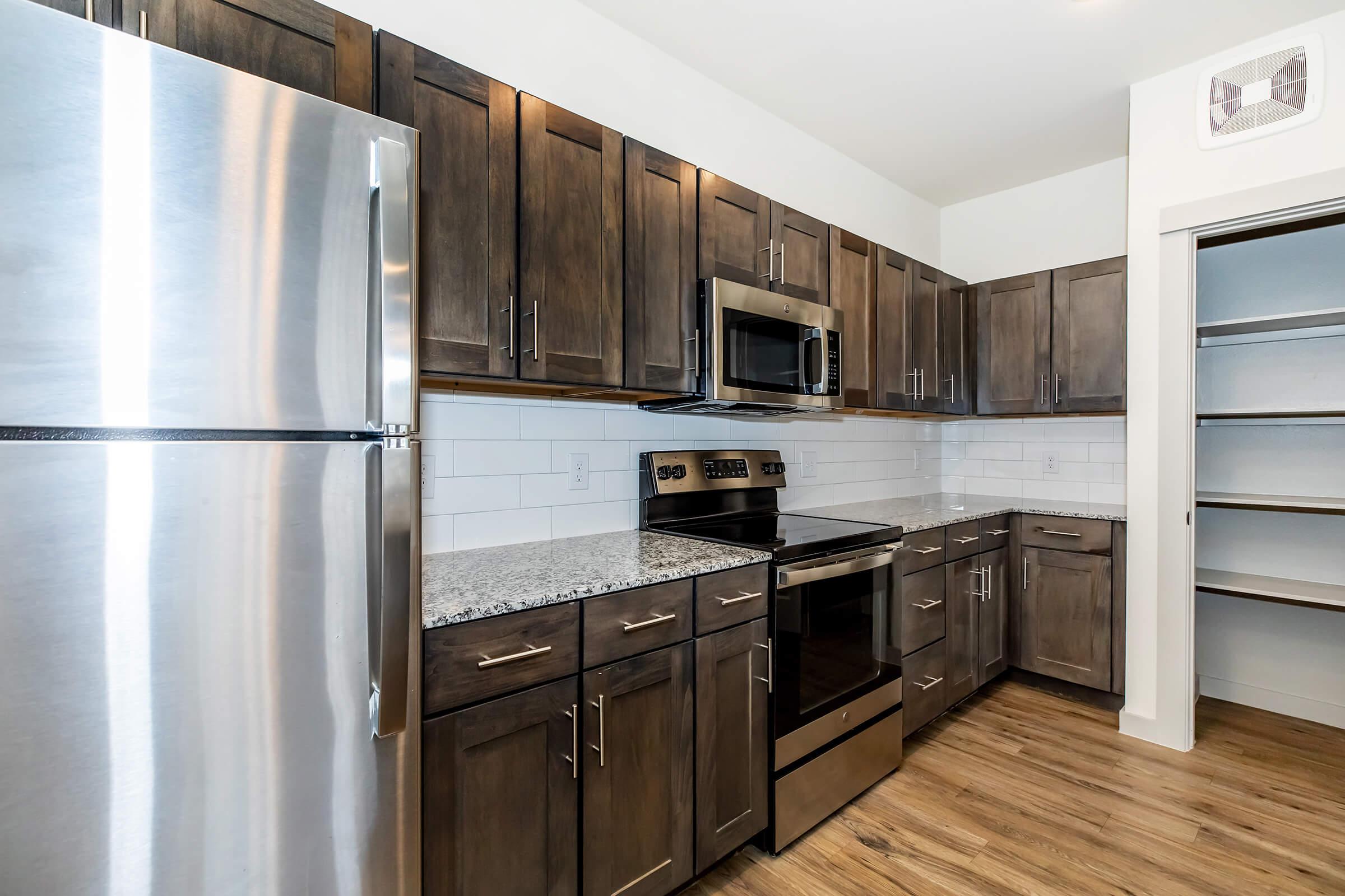 Modern kitchen featuring dark wooden cabinetry, stainless steel appliances including a refrigerator and microwave, and a granite countertop. The space has white tiles as a backsplash and wooden flooring, creating a sleek and stylish look. A pantry is visible in the background.