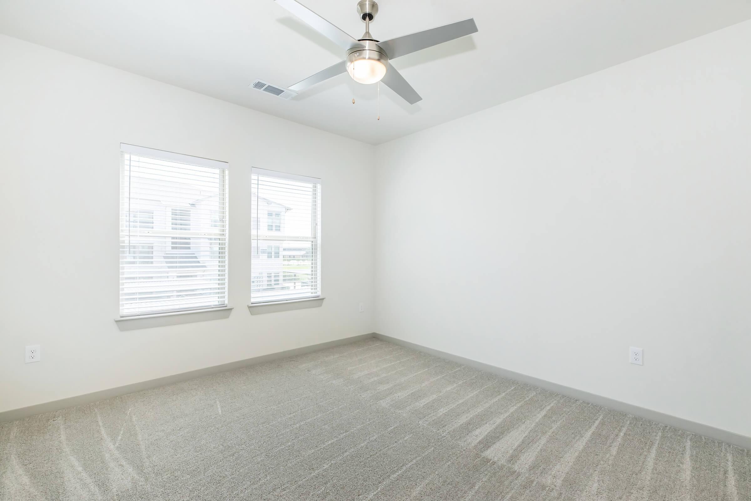 A spacious, empty bedroom featuring light-colored walls, a ceiling fan with a light fixture, and two windows with white blinds. The carpeted floor has a subtle pattern, and the room is well-lit with natural light coming through the windows.