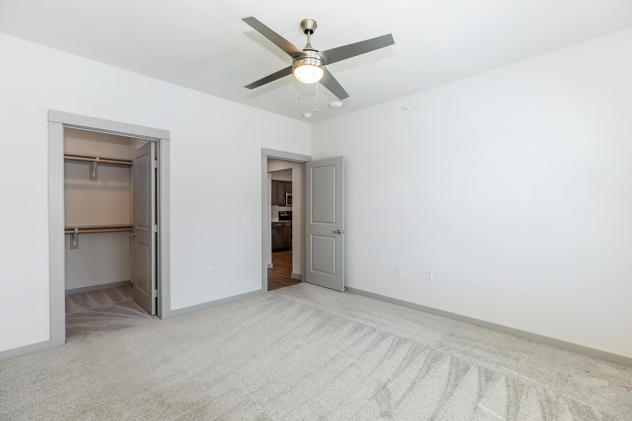 A spacious, well-lit bedroom featuring a ceiling fan, light-colored walls, and plush carpet. On one side, there is an open closet with shelving, while the door on the left leads to another room. The room appears modern and inviting, suitable for a comfortable living space.