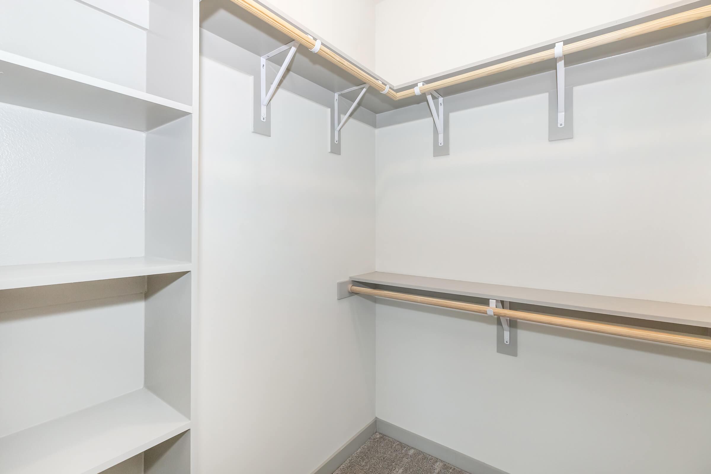 Empty closet with light gray walls, featuring a shelf on the left side and two hanging rods along the back wall. The floor is carpeted in a neutral gray tone.