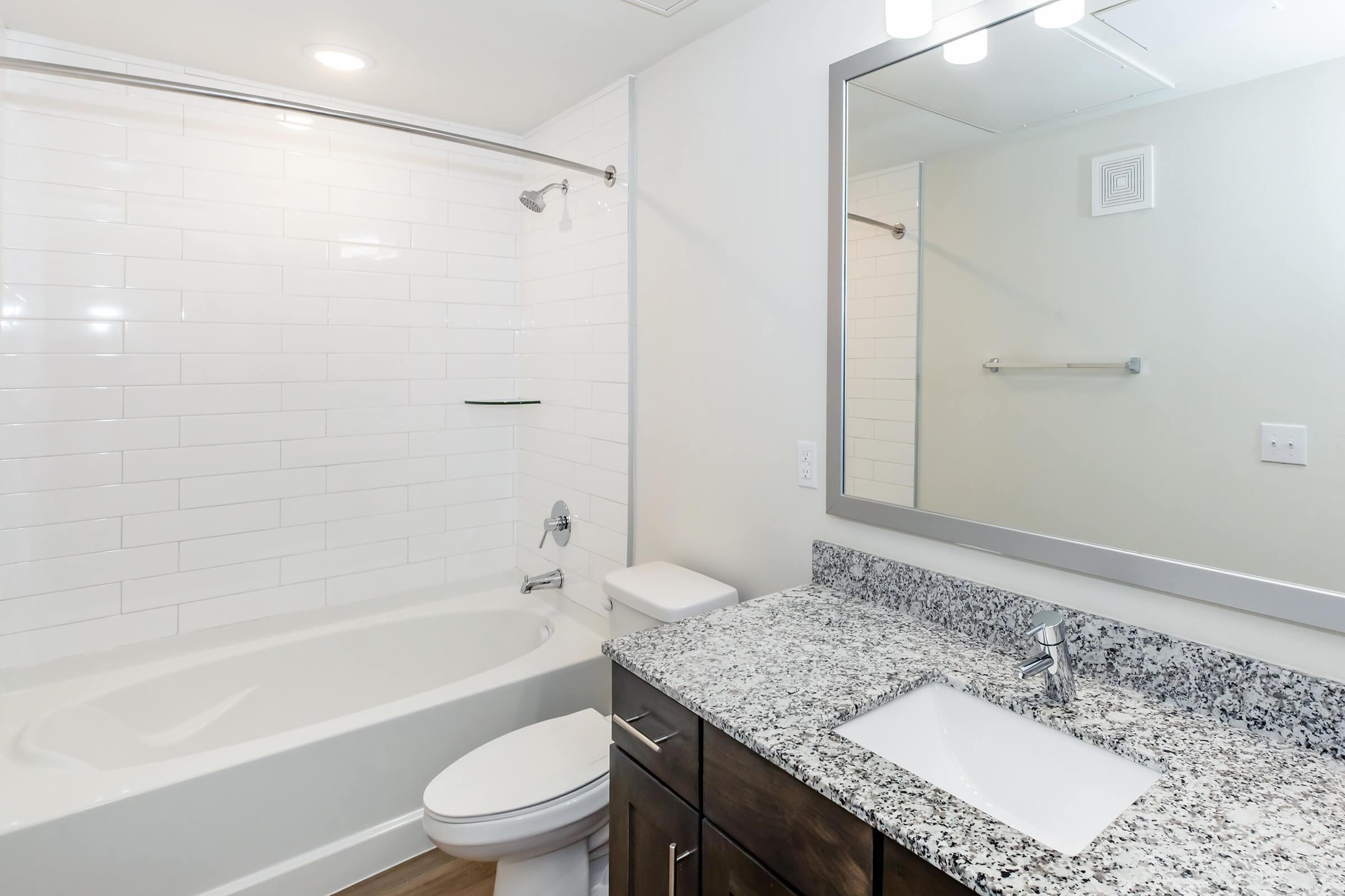 A modern bathroom featuring a white tiled shower and bathtub, a dark wood vanity with a granite countertop, a sleek sink, and a large mirror with built-in lighting. The space is well-lit and has a clean, minimalist design.