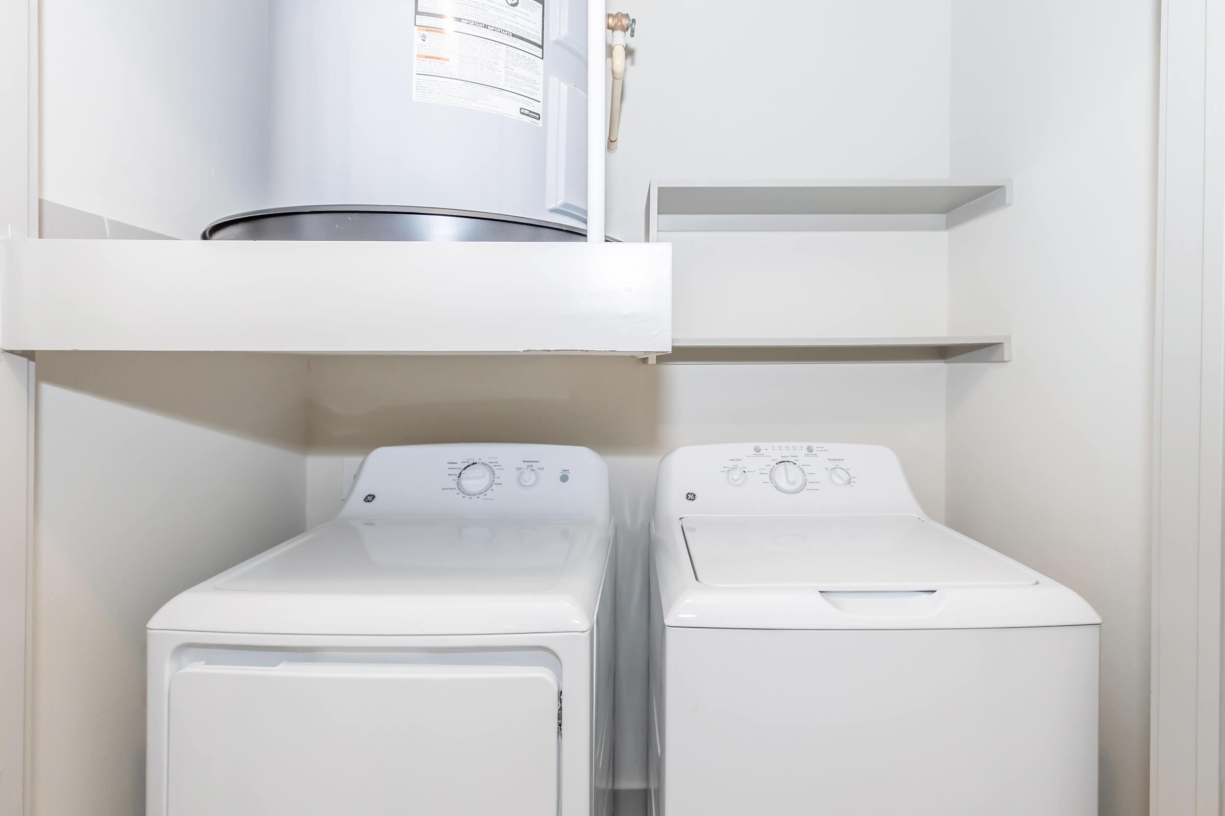 A laundry room featuring a white washing machine and dryer side by side, with a hot water heater above them. There are also two empty shelves mounted on the wall for storage. The walls are painted a light color, creating a clean and organized appearance.