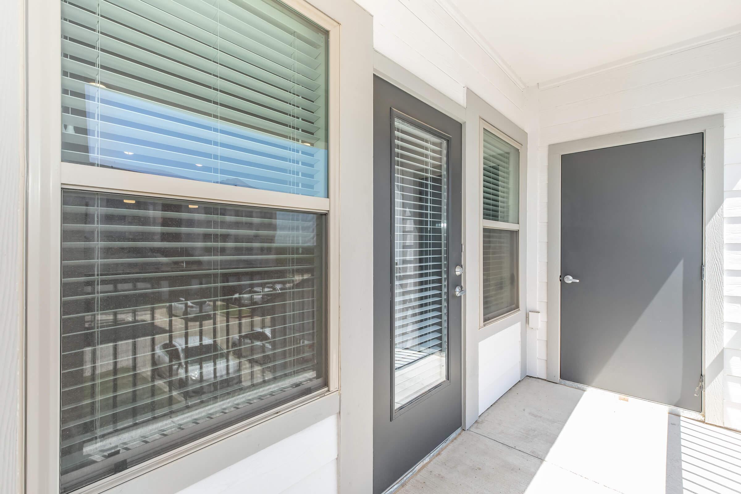 A well-lit entrance area featuring two large windows with blinds and a dark gray door. The walls are painted white, and the concrete floor is clean. The scene includes a glimpse of what appears to be a courtyard or outdoor area through the windows.