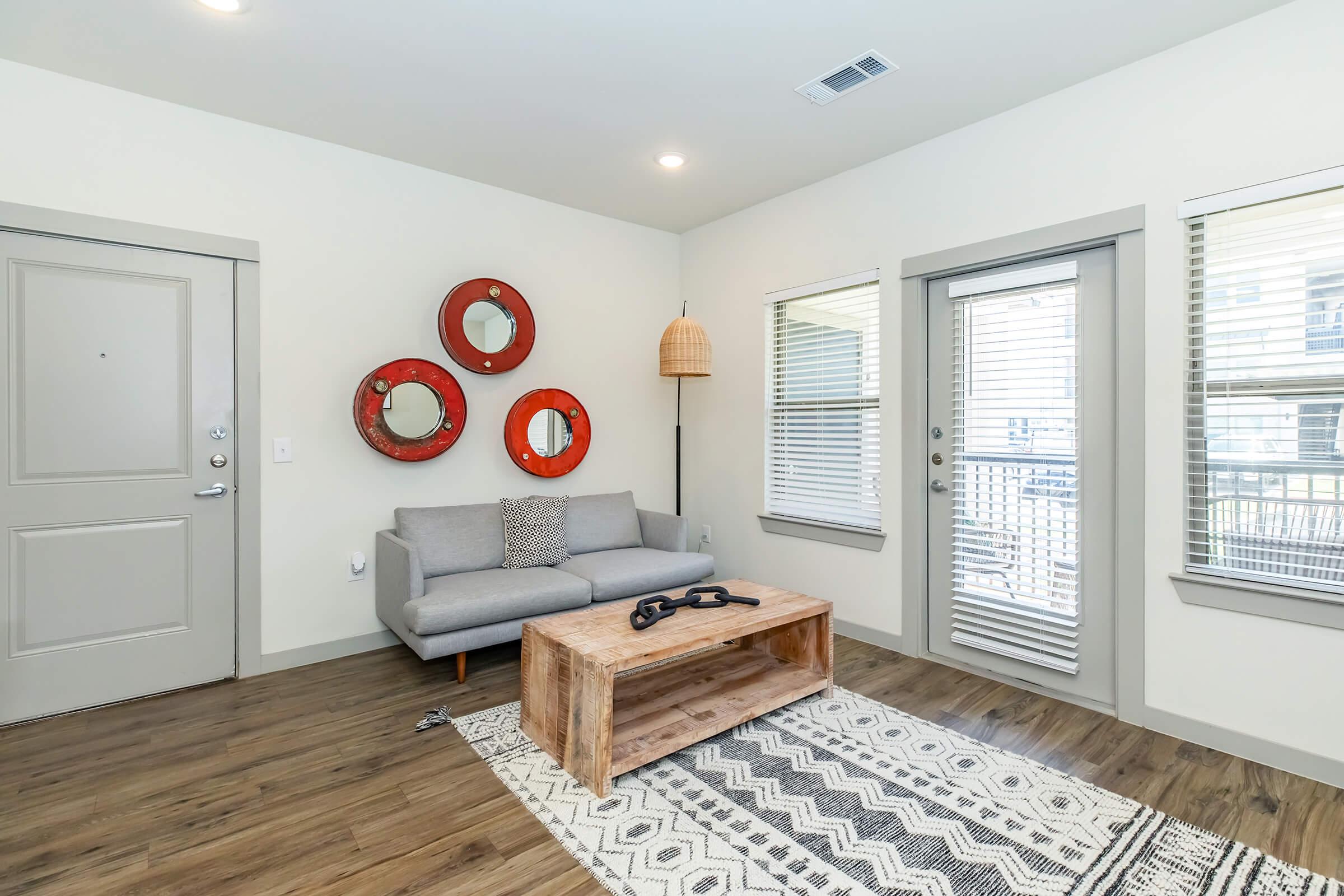 A modern living room featuring a gray sofa with decorative pillows, a wooden coffee table, and a patterned rug. The wall is adorned with three circular mirrors with red frames. There are large windows with blinds and a door leading outside. Warm, neutral tones create a cozy atmosphere.