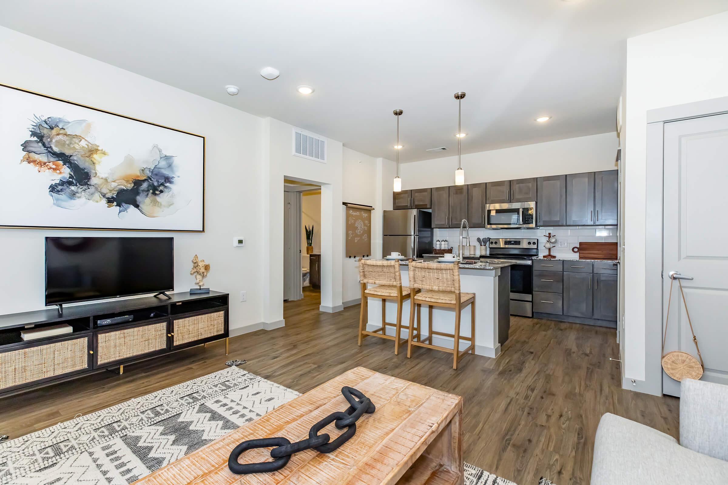 Interior of a modern apartment featuring a stylish living area with a flat-screen TV and an abstract wall art piece. A kitchen with stainless steel appliances and a dining area with bar stools is visible. The flooring is wood, and the decor is contemporary, including a decorative rug and a wooden coffee table.