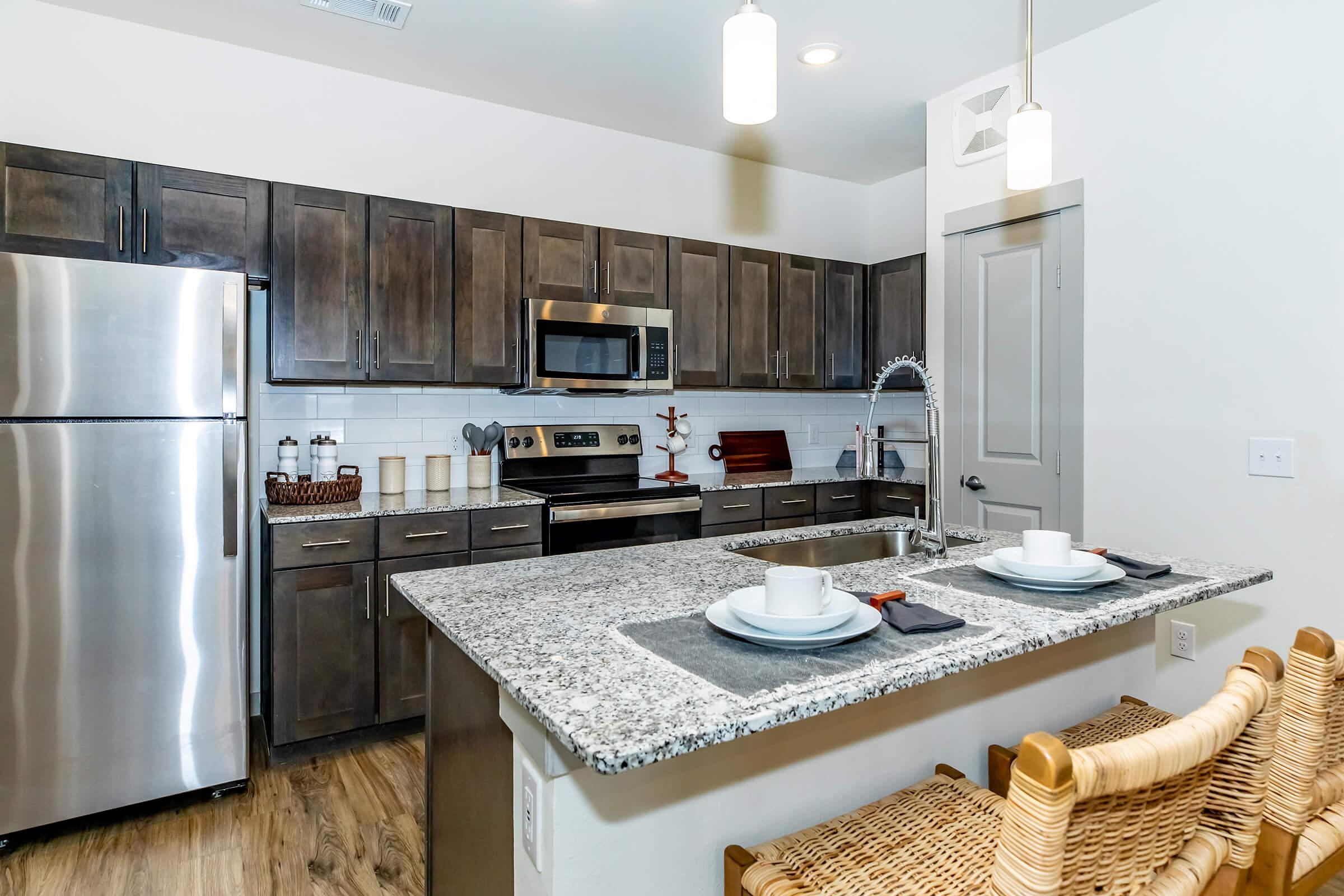 Modern kitchen featuring dark wooden cabinetry, stainless steel appliances, a granite countertop island with two place settings, and woven barstools. The space is well-lit with pendant lights and has a clean, contemporary design, complemented by a tiled backsplash and wood flooring.