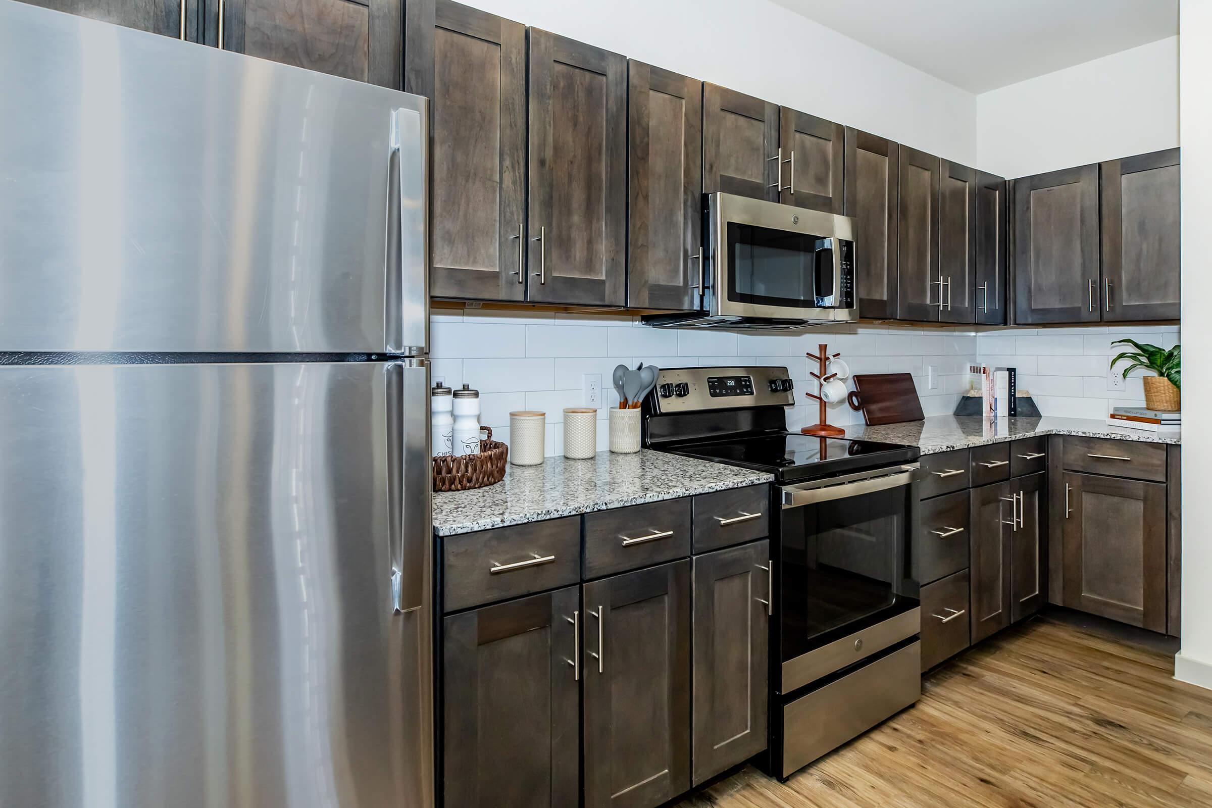 A modern kitchen featuring stainless steel appliances, including a refrigerator and an oven, with dark wooden cabinets and a granite countertop. The space is well-lit, showcasing a tidy arrangement of jars and utensils on the counter, along with a potted plant for added decor.