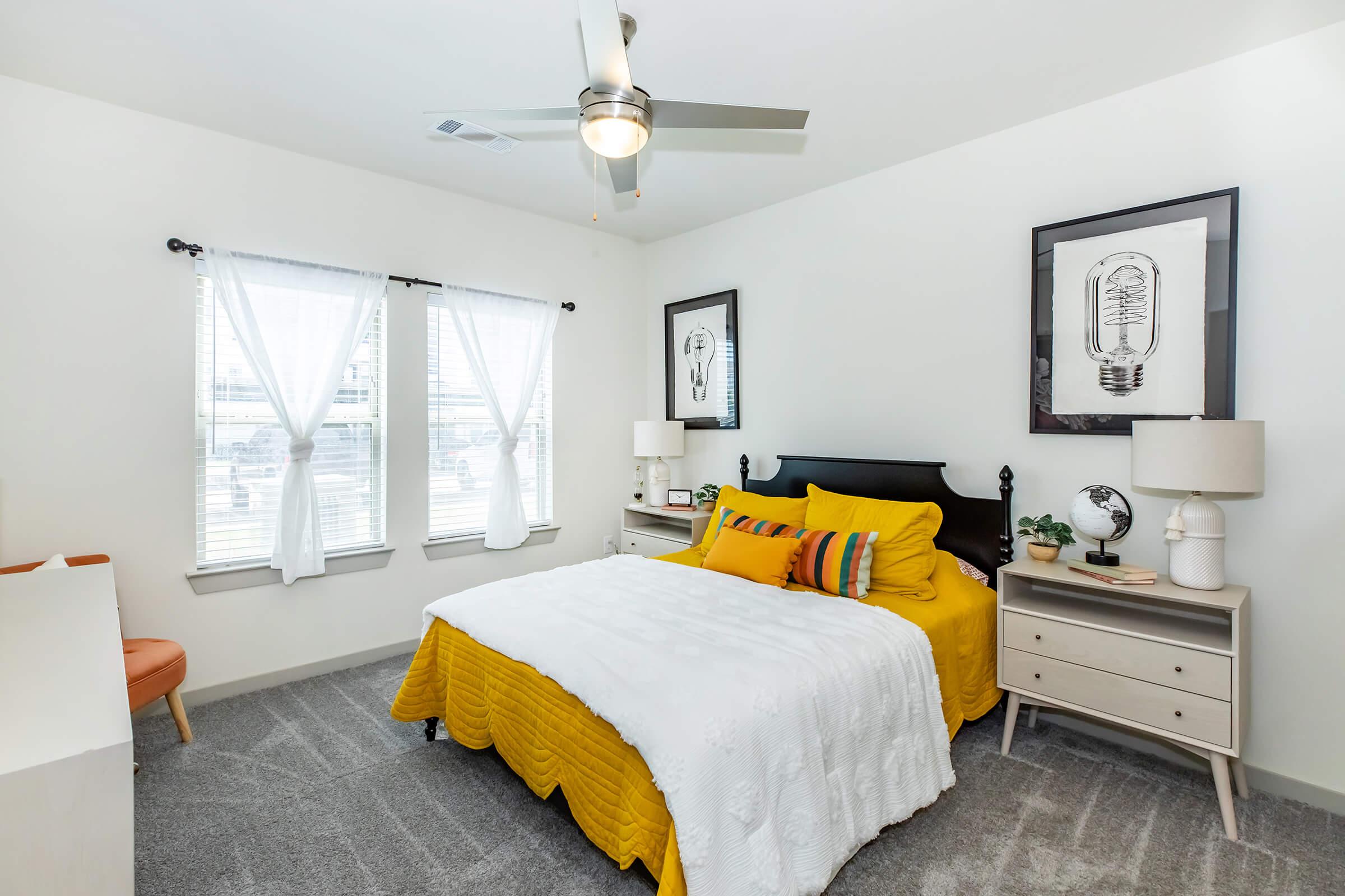 A cozy bedroom featuring a queen-sized bed with yellow and white bedding, two framed art pieces on the wall, and a bedside table with lamps. The room has large windows with sheer curtains, light gray carpet, and a chair in the corner, creating a bright and inviting atmosphere.