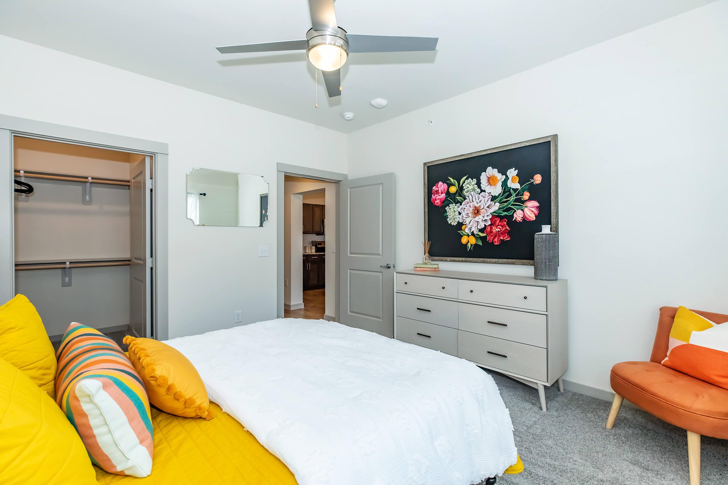 A cozy bedroom featuring a white bed with yellow and striped pillows, a large floral artwork on the wall, a dresser, and an armchair. There’s a closet with an open door, a ceiling fan, and light-colored walls, creating a bright and inviting atmosphere.