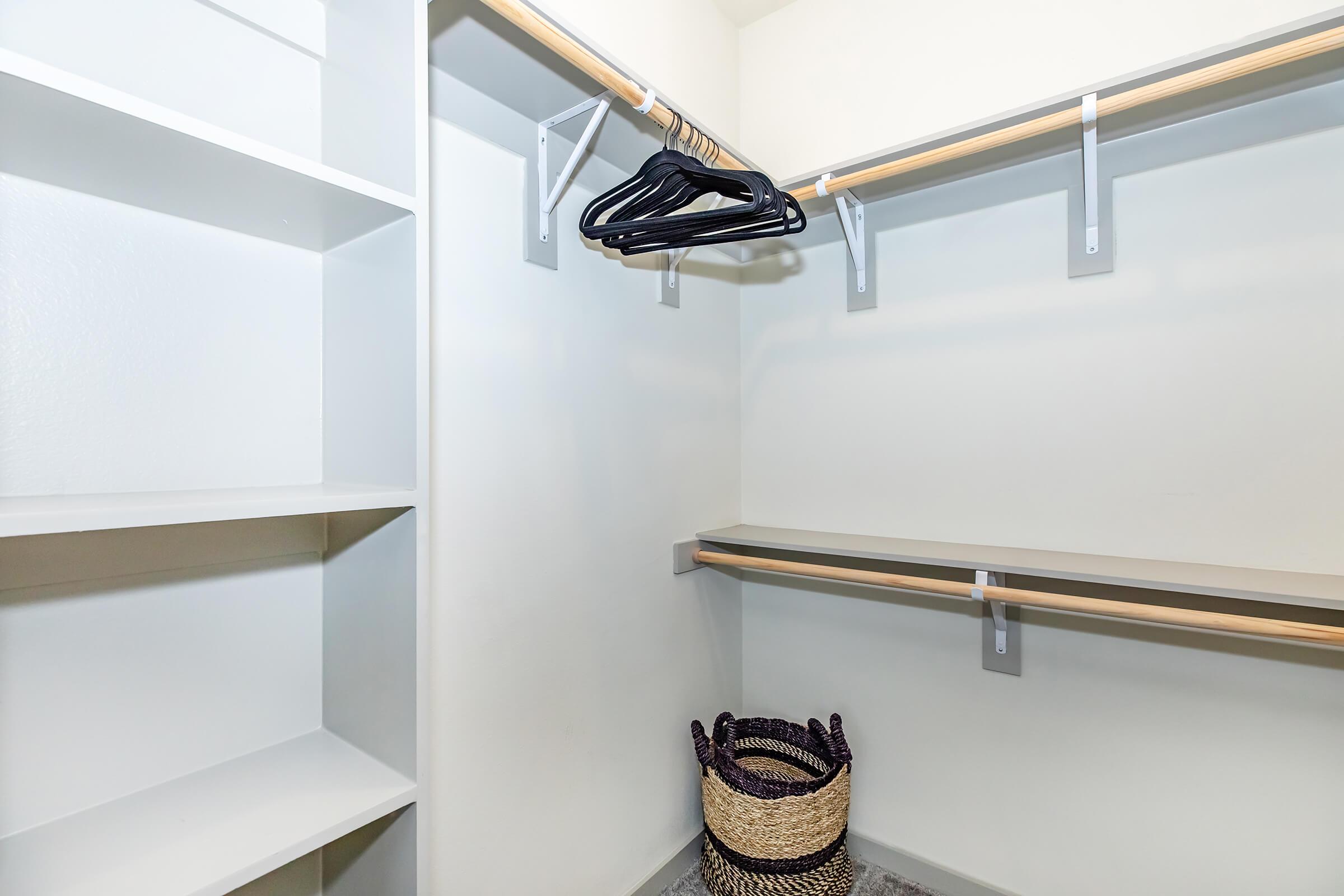 A clean, empty closet with white shelves and a wooden hanging rod. There are a few black hangers hanging from the rod and a woven basket on the floor. The walls are light-colored, creating a bright and spacious feel.