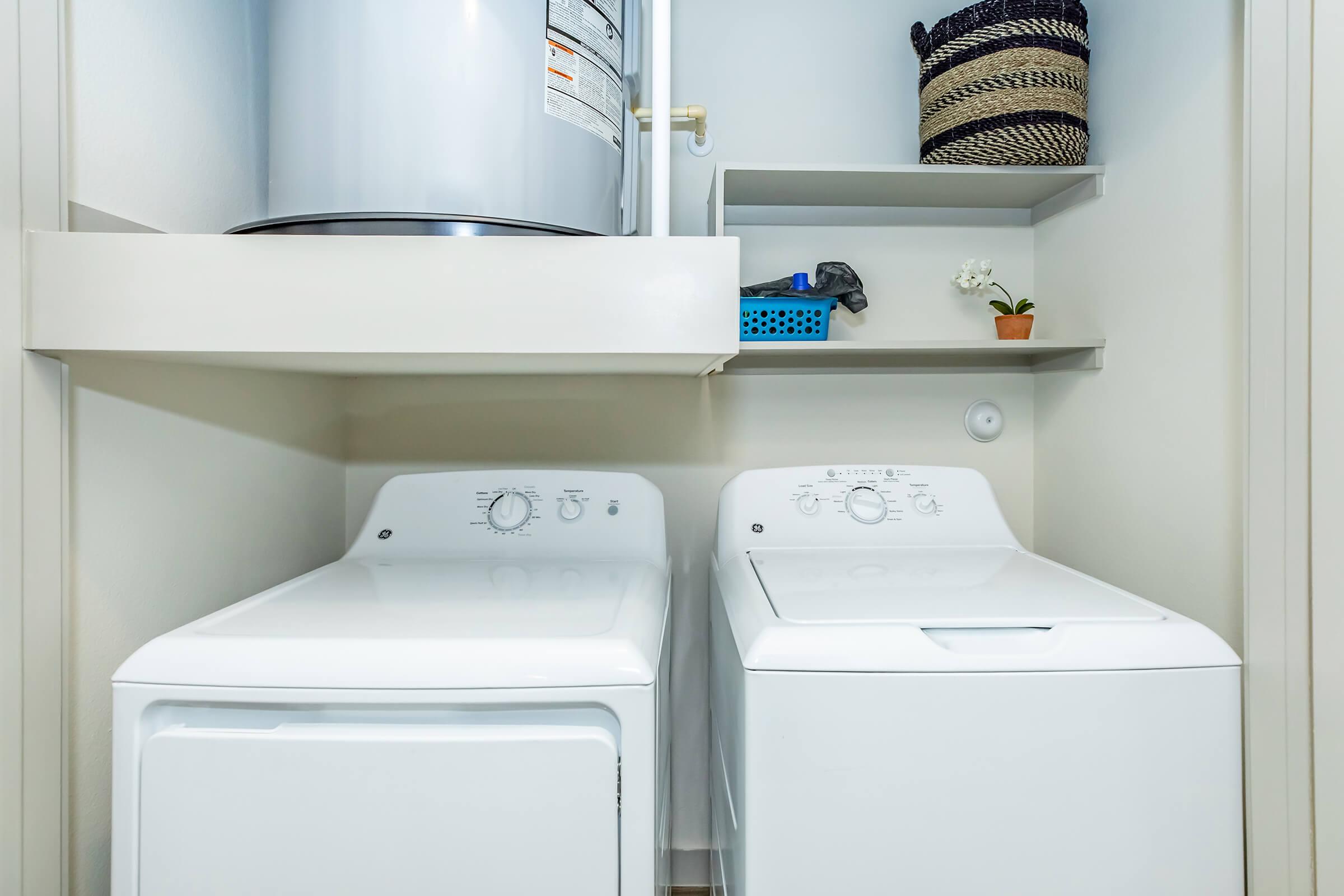 A bright laundry room featuring a stackable washer and dryer set in white. Above the appliances, there is a round water heater. The shelves on the wall hold a small plant and a striped basket, adding a touch of decor to the space. The overall look is clean and organized.