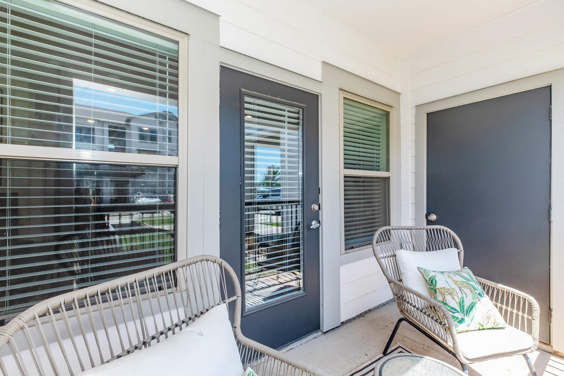 A cozy outdoor patio area featuring two chairs with cushions and a small table. The space has large windows with blinds, providing natural light. There are two gray doors leading into an enclosed area, creating a welcoming atmosphere for relaxation.