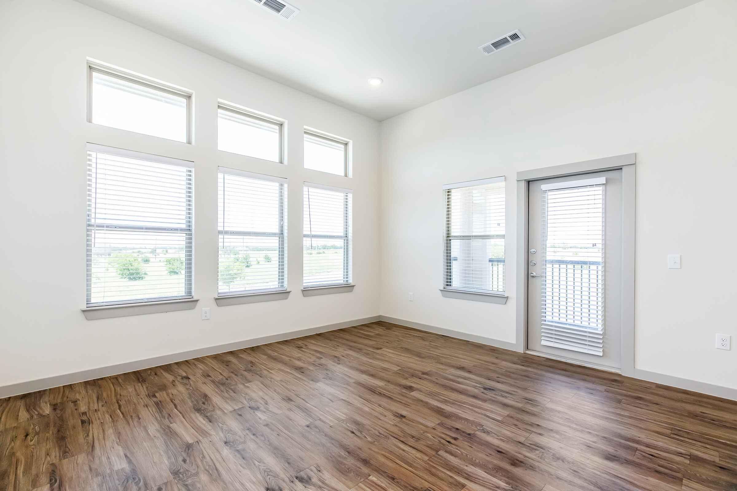 Bright and spacious interior of a vacant room featuring large windows with blinds, allowing natural light to fill the space. The wooden floor adds warmth, and there is a door leading outside. The walls are painted in a light color, enhancing the room's airy feel.