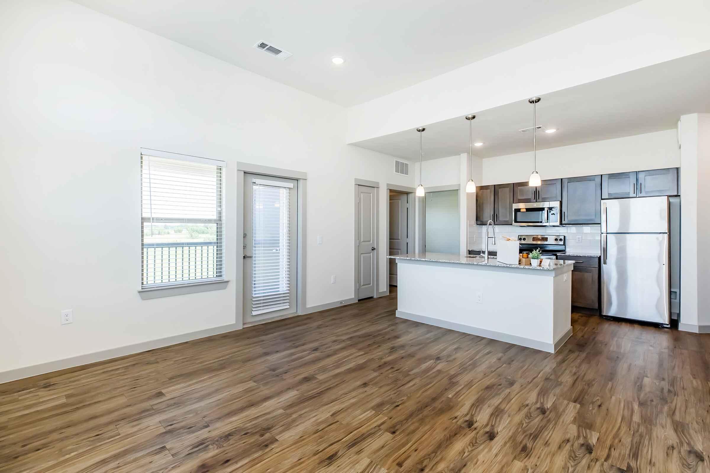 A modern, open-concept kitchen and living area featuring wooden flooring, stainless steel appliances, and pendant lighting. Large windows provide natural light, and a sliding glass door leads to an outdoor space. The kitchen has a central island with bar seating.