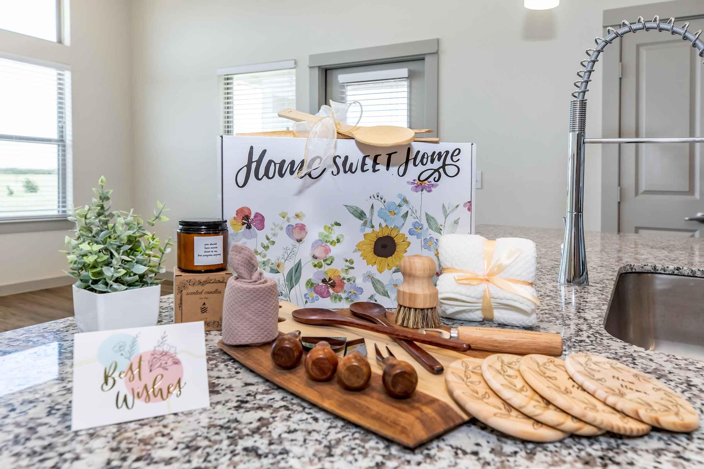 A beautifully arranged kitchen gift set featuring a "House Sweet Home" box, decorative flowers, kitchen utensils like wooden spoons and a rolling pin, a candle, dish towels, and a greeting card that says "Best Wishes," all displayed on a granite countertop with a plant in the background.