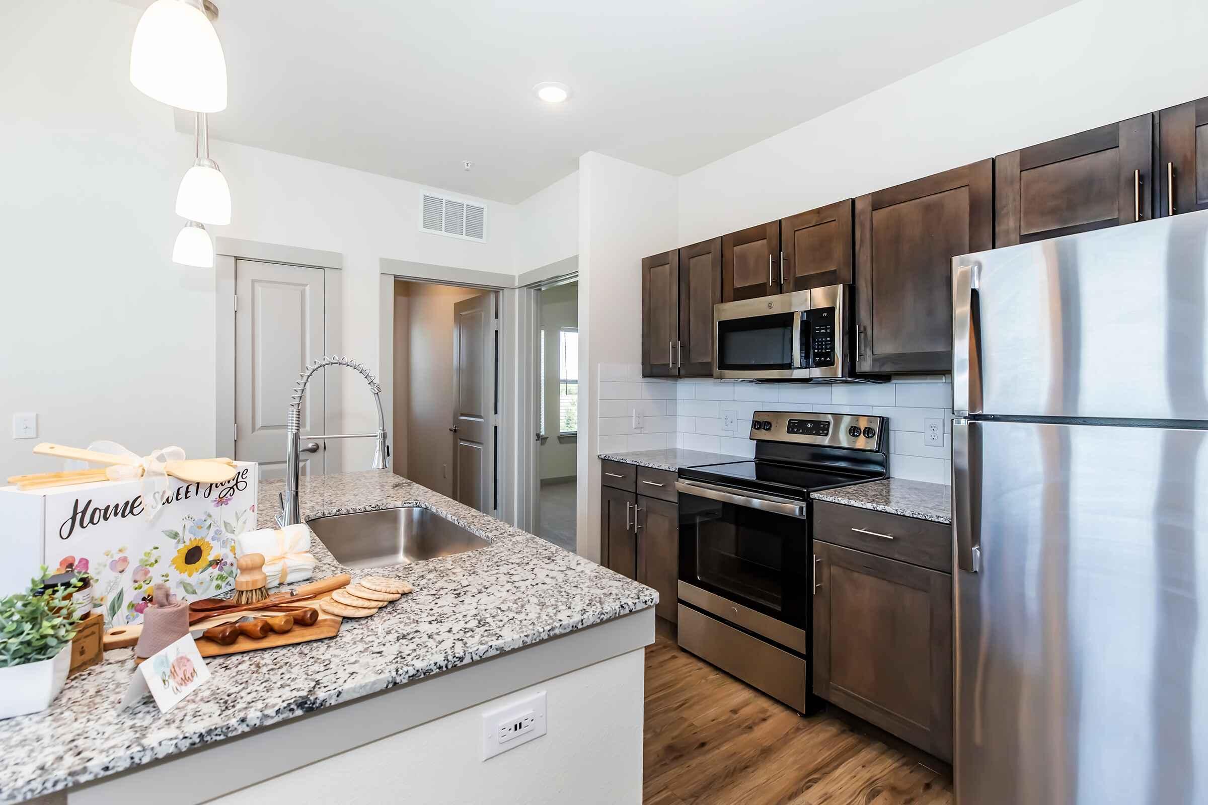 A modern kitchen featuring dark wooden cabinets, stainless steel appliances, and a large granite countertop. The space includes a sink with a sleek faucet, a stove, and an oven, along with a decorative wooden serving tray on the countertop. Bright lighting and a window suggest a spacious, airy environment.
