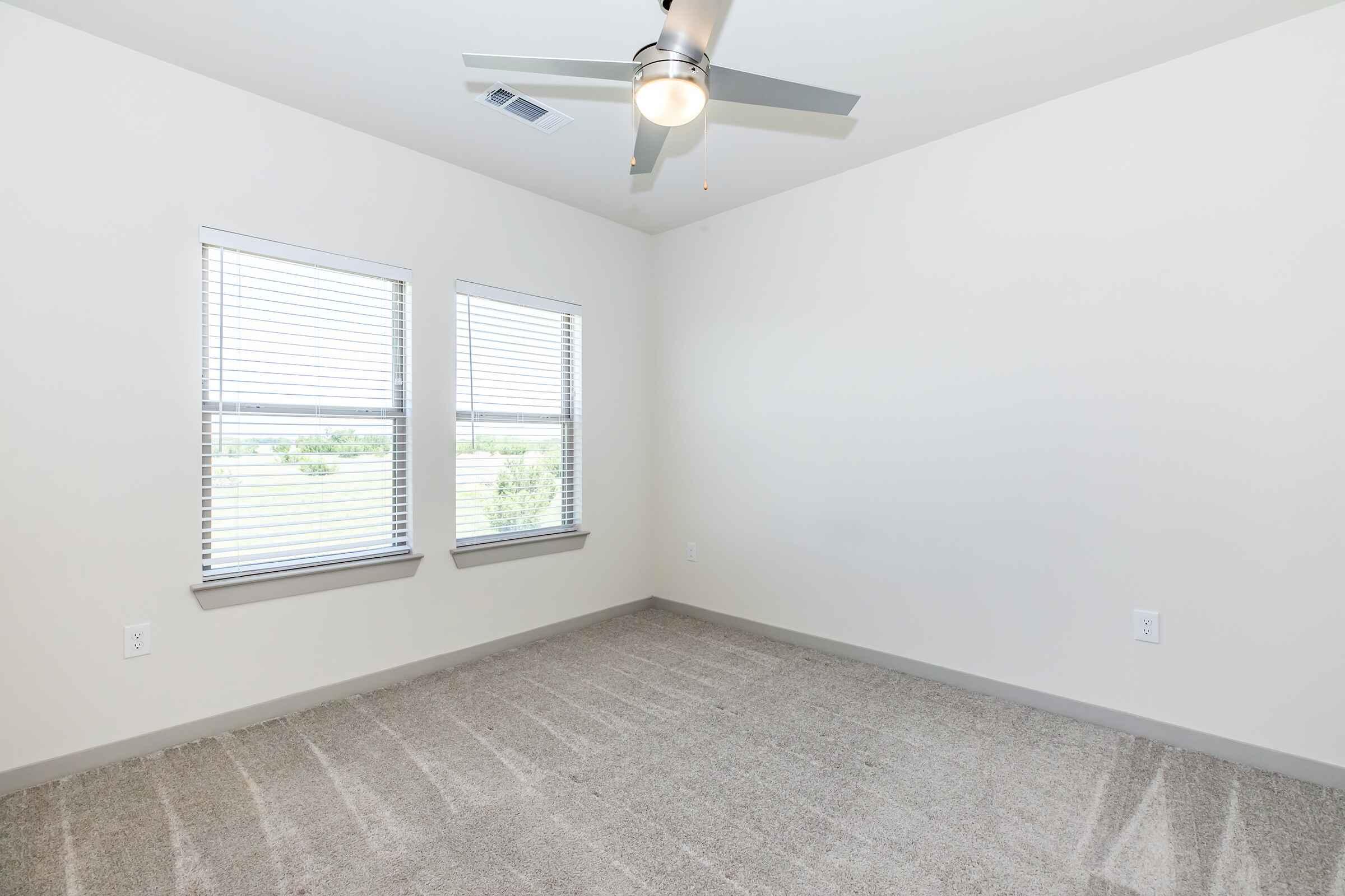 Interior view of an empty room with light-colored walls and carpeted flooring. Two windows with blinds allow natural light to enter. A ceiling fan is mounted on the ceiling, adding a modern touch. The room appears spacious and is ready for furnishings.