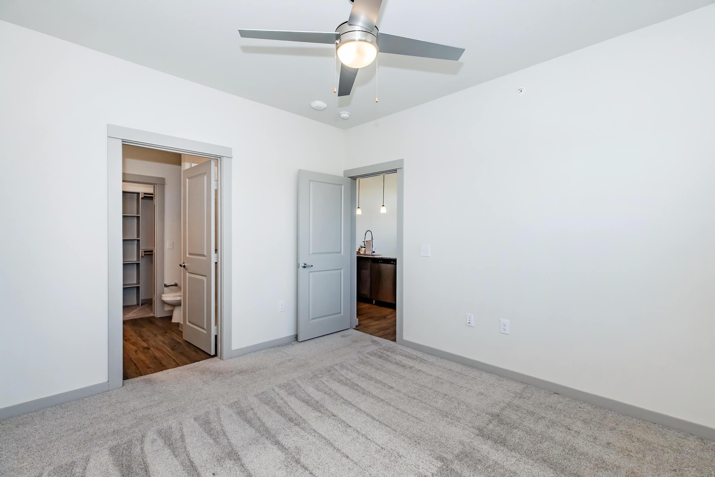 A spacious, minimally furnished bedroom featuring light gray walls and a ceiling fan. It has plush carpet flooring and doors leading to a bathroom on the left and a kitchen or dining area on the right. The lighting is bright, highlighting the room's open layout.