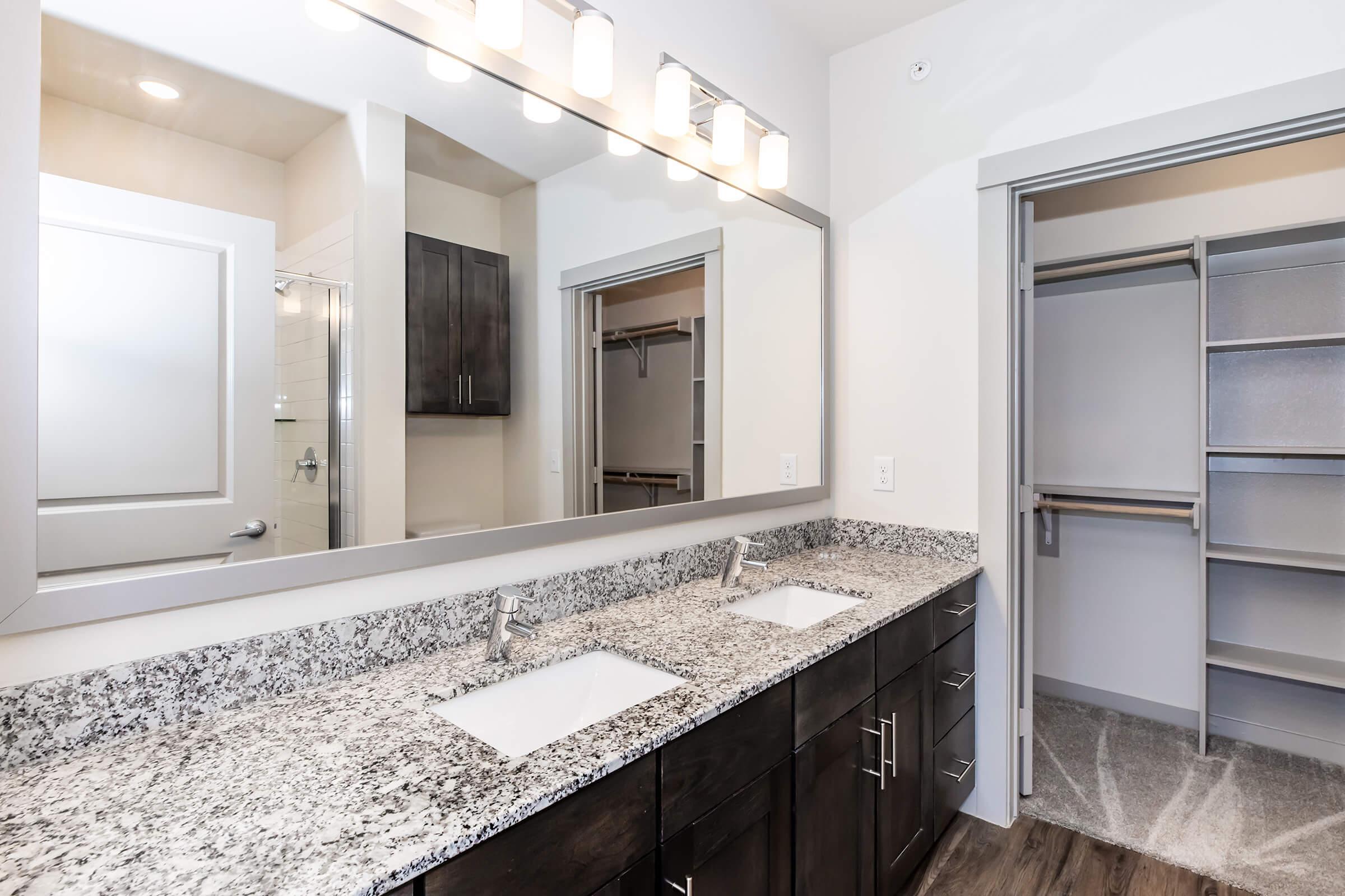 A modern bathroom featuring a double vanity with two sinks and a large mirror above, complemented by stylish light fixtures. The countertop is made of speckled gray granite. To the right, there is an open closet with shelving and hanging space, showcasing a clean, minimalist design.