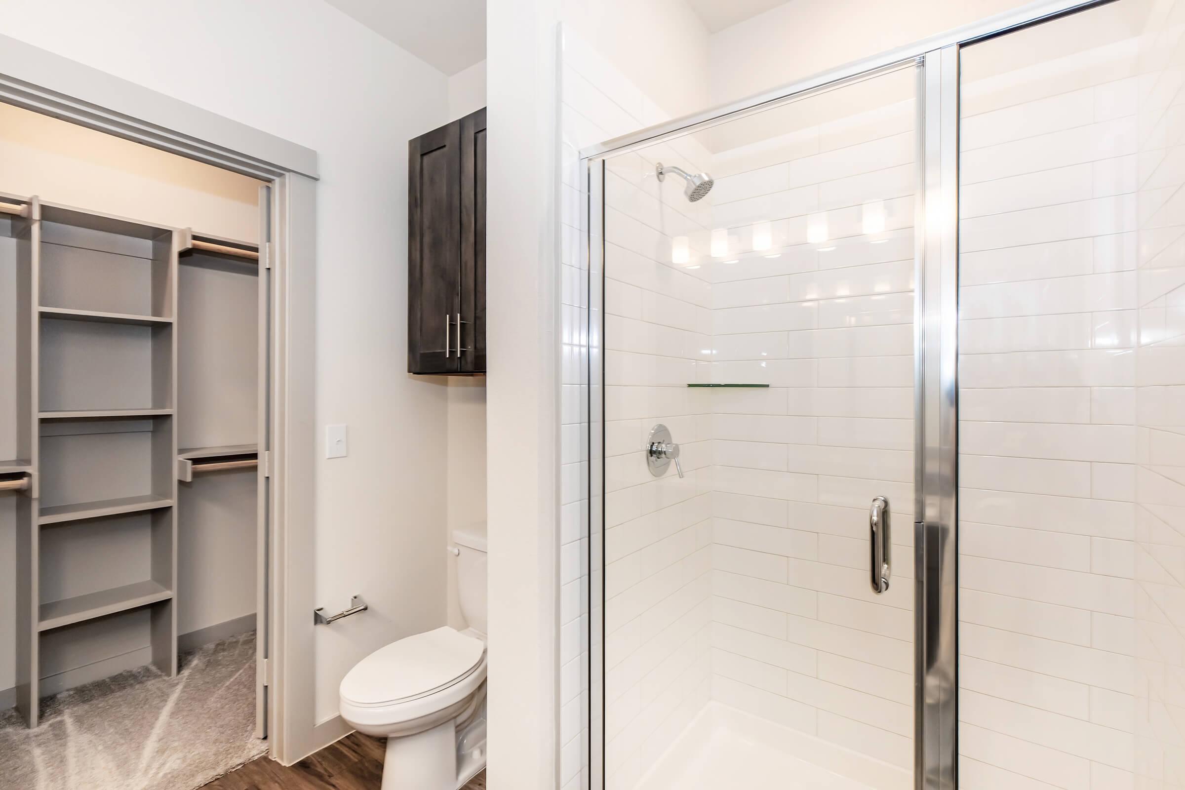 A modern bathroom featuring a glass shower enclosure with white tile, a toilet, and a large walk-in closet with shelving visible through an open doorway. The space has neutral colors and contemporary fixtures, creating a clean and bright aesthetic.