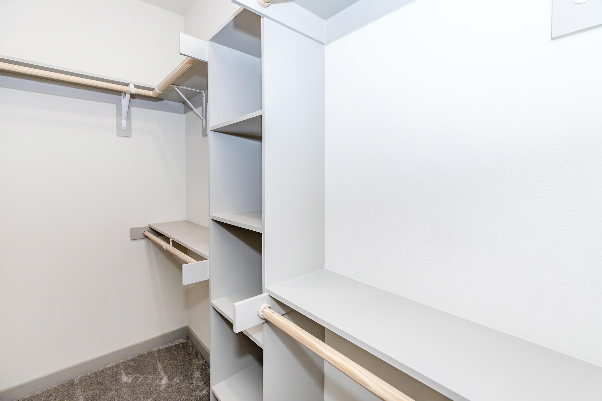 A spacious and organized walk-in closet featuring multiple shelves and hanging rods. The closet has a neutral color scheme with light gray walls and wooden clothing rods. The floor is carpeted, providing a cozy feel to the storage area.