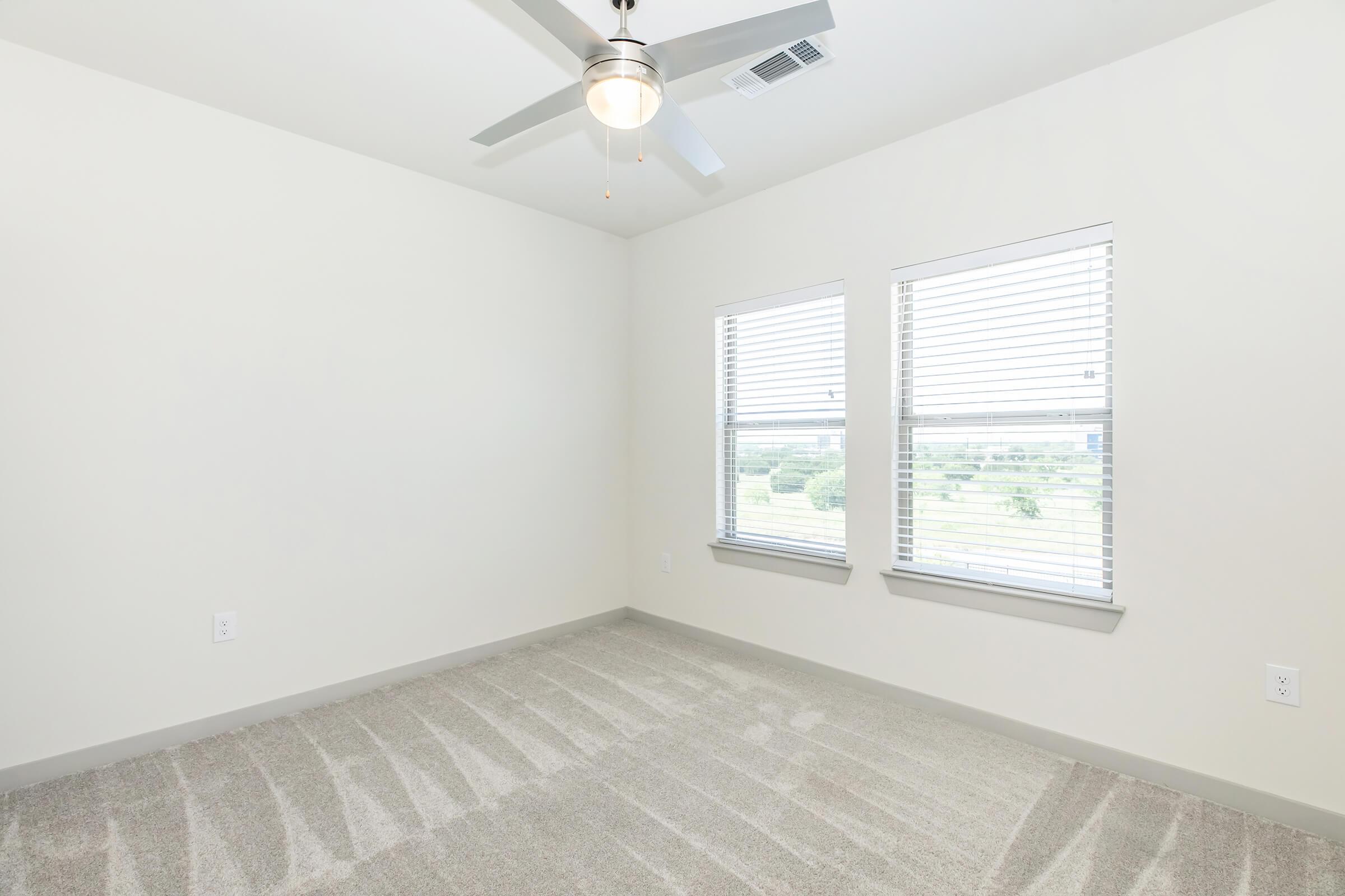 Empty room with light-colored walls and a ceiling fan. Two windows with white blinds allow natural light to fill the space. The floor is covered with light gray carpet, and the room has a minimalist design, making it feel spacious and bright.