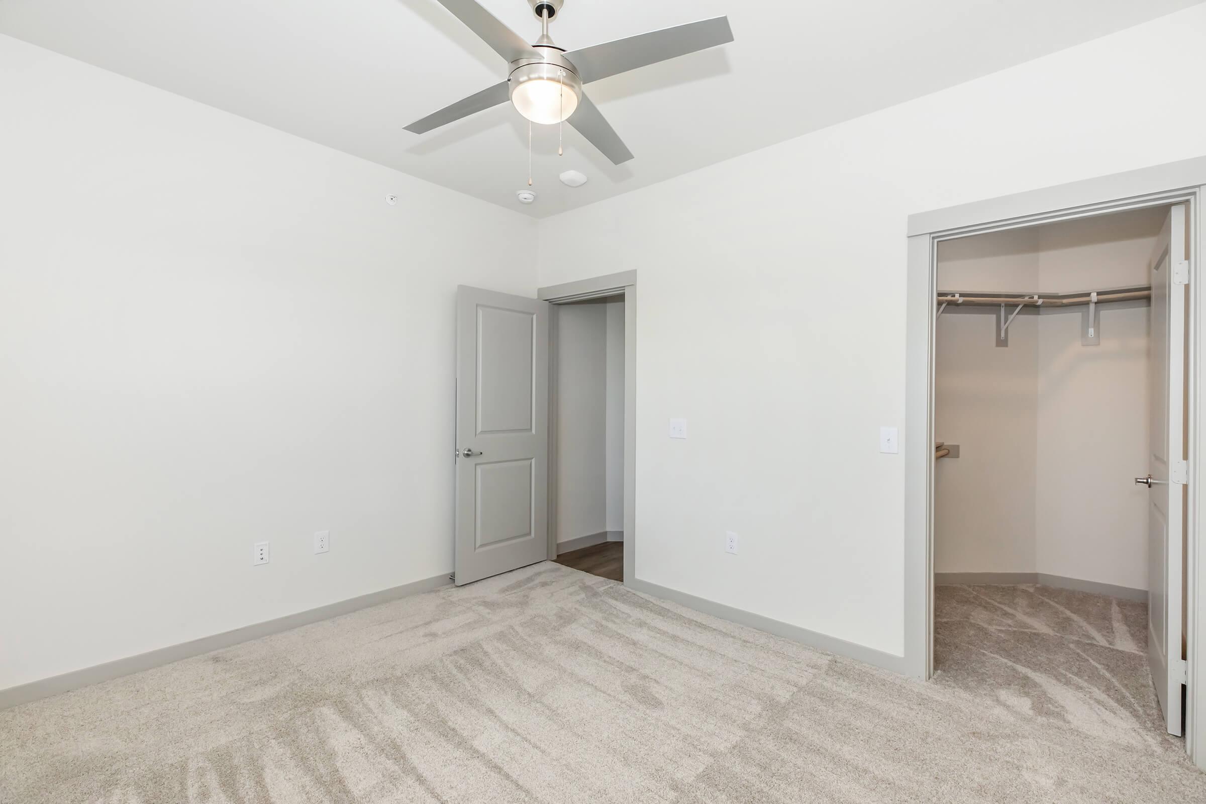 A minimalistic bedroom featuring light-colored walls, a ceiling fan, and plush carpeting. There is a closet with sliding doors to the right, and the room is brightly lit, creating a spacious and inviting atmosphere. The design is contemporary, with clean lines and neutral tones.