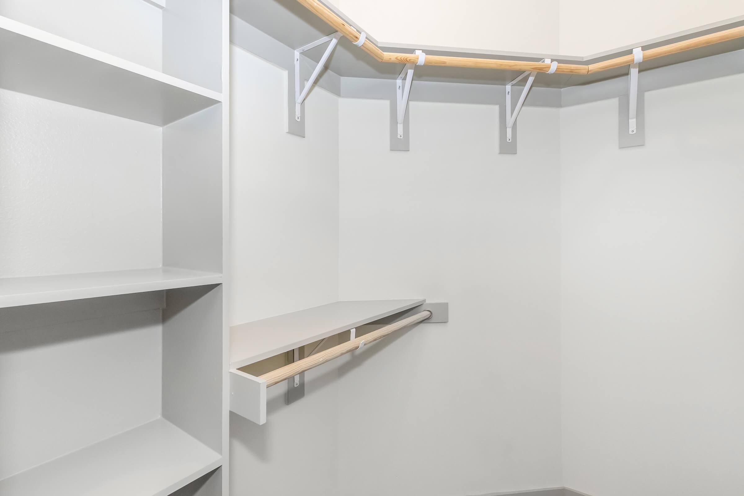 An empty closet with light gray walls, featuring shelves on the left side and a hanging rod with hooks on the right. There is an open space for hanging clothes and a shelf above the hanging area, creating a clean and minimalist storage solution.