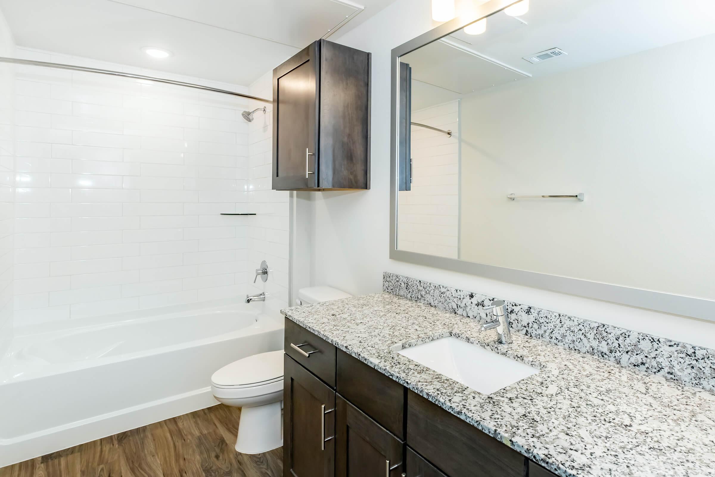A modern bathroom featuring a white tiled shower with a glass door, a bathtub, a dark wooden vanity with a speckled granite countertop, a rectangular sink, and a large wall mirror. The floor is made of wooden planks, and the walls are painted in a light color.
