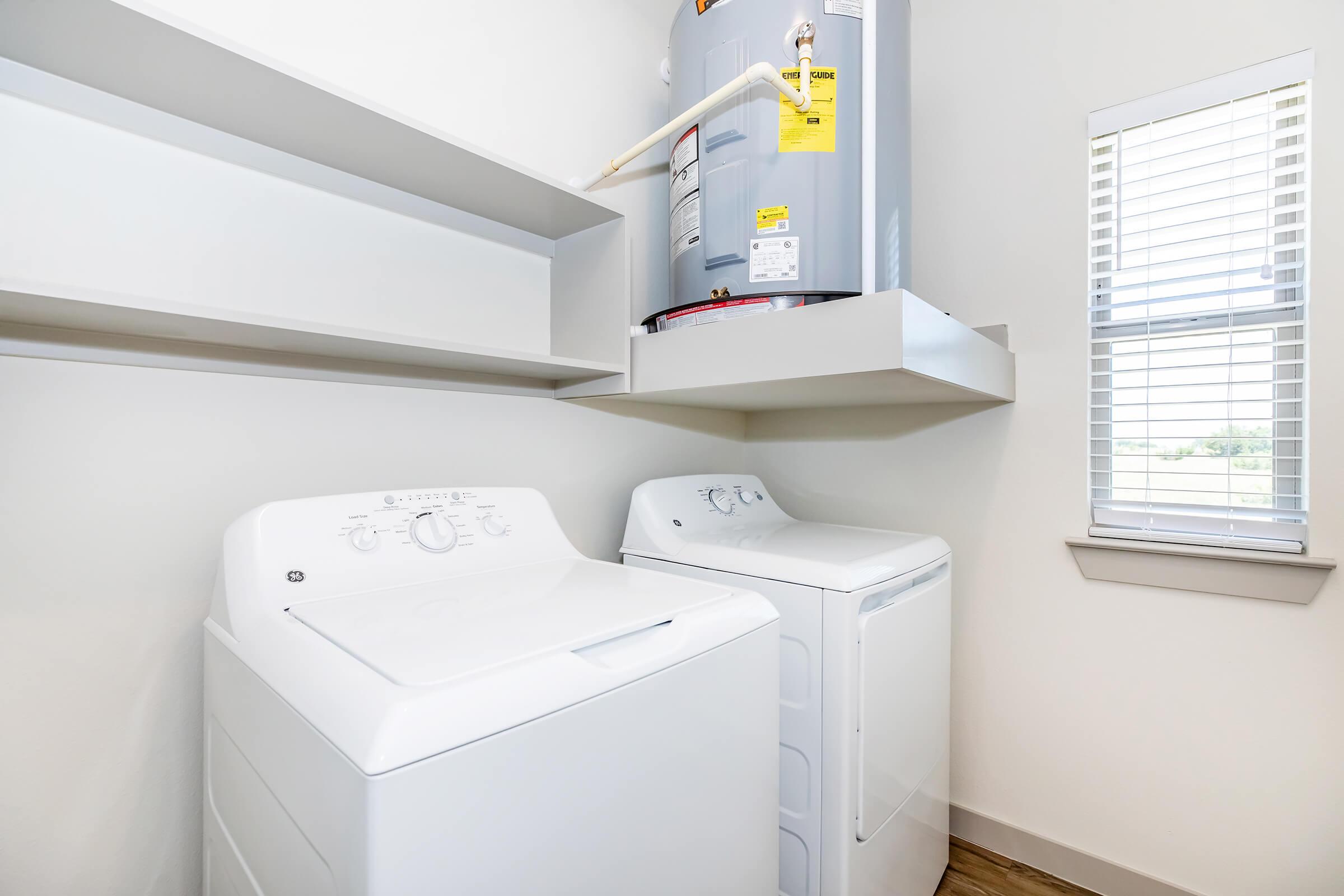 A modern laundry room featuring a white washing machine and dryer side by side. Above the machines is a shelf and a water heater mounted on the wall. The room has light-colored walls and a window with blinds, allowing natural light to enter.