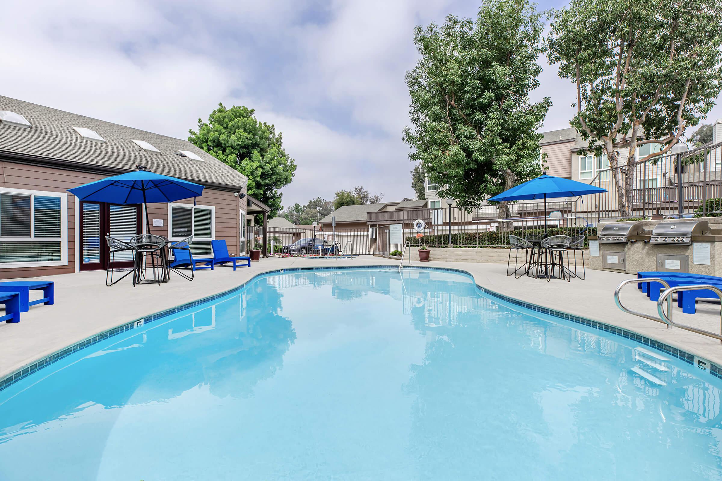 Two Bedroom luxury Apartments in Riverside CA - Canyon Crest Views - Resort Style Swimming Pool With Blue Umbrellas, Lounge Chairs, Patio Tables, And Nearby Barbecue Grills