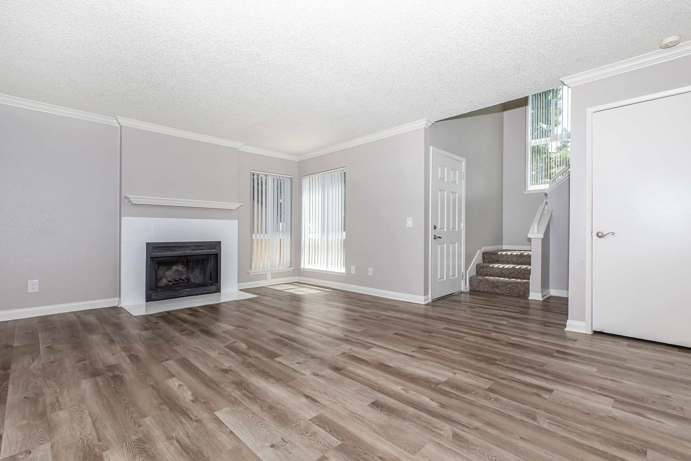 One Bedroom luxury Apartments in Riverside CA - Canyon Crest Views - Open Concept Living Room With Wood Style Flooring, Corner Fireplace, Staircase Entry, And Tall Windows With Vertical Blinds