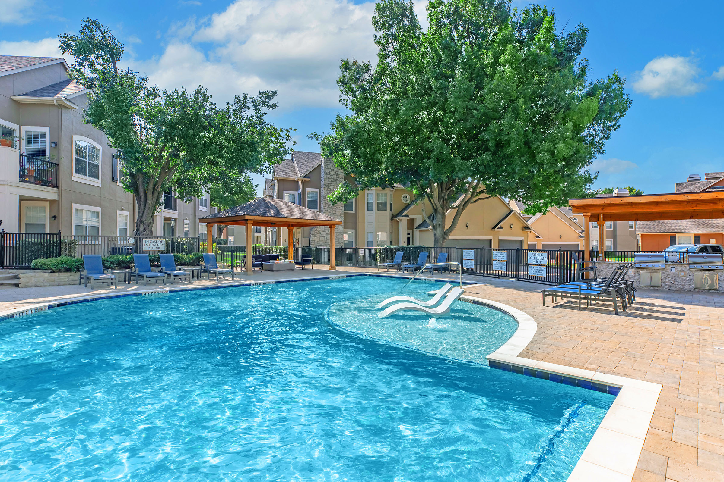 A bright and inviting outdoor pool area surrounded by lounge chairs and shaded cabanas. The pool features a gentle curve, with steps leading in, and is bordered by lush greenery and nearby apartment buildings, all under a clear blue sky.