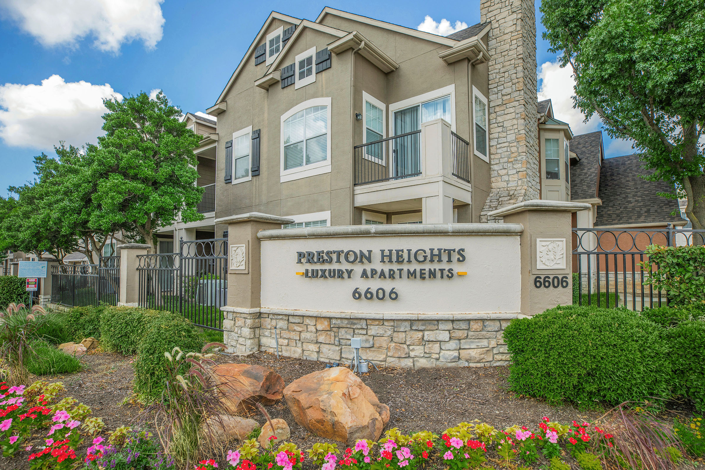 Exterior view of Preston Heights Luxury Apartments, featuring a well-maintained stone entrance sign with "Preston Heights Luxury Apartments" and the number "6606." The building has multiple stories with balconies, surrounded by lush green trees and colorful flower beds in the foreground.