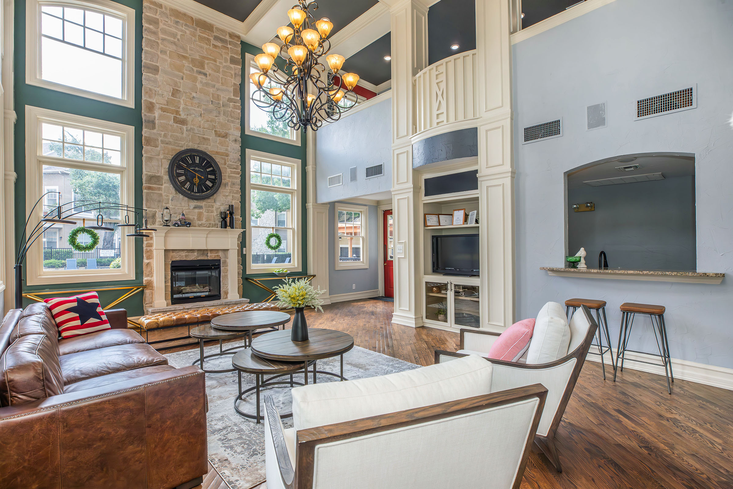 A spacious living room featuring a high ceiling with a chandelier, large windows, and a stone fireplace. The area is furnished with a brown leather sofa, white chairs, and round coffee tables. Decorative elements include a wall clock and green wreaths on the windows, with a modern bar counter in the background.