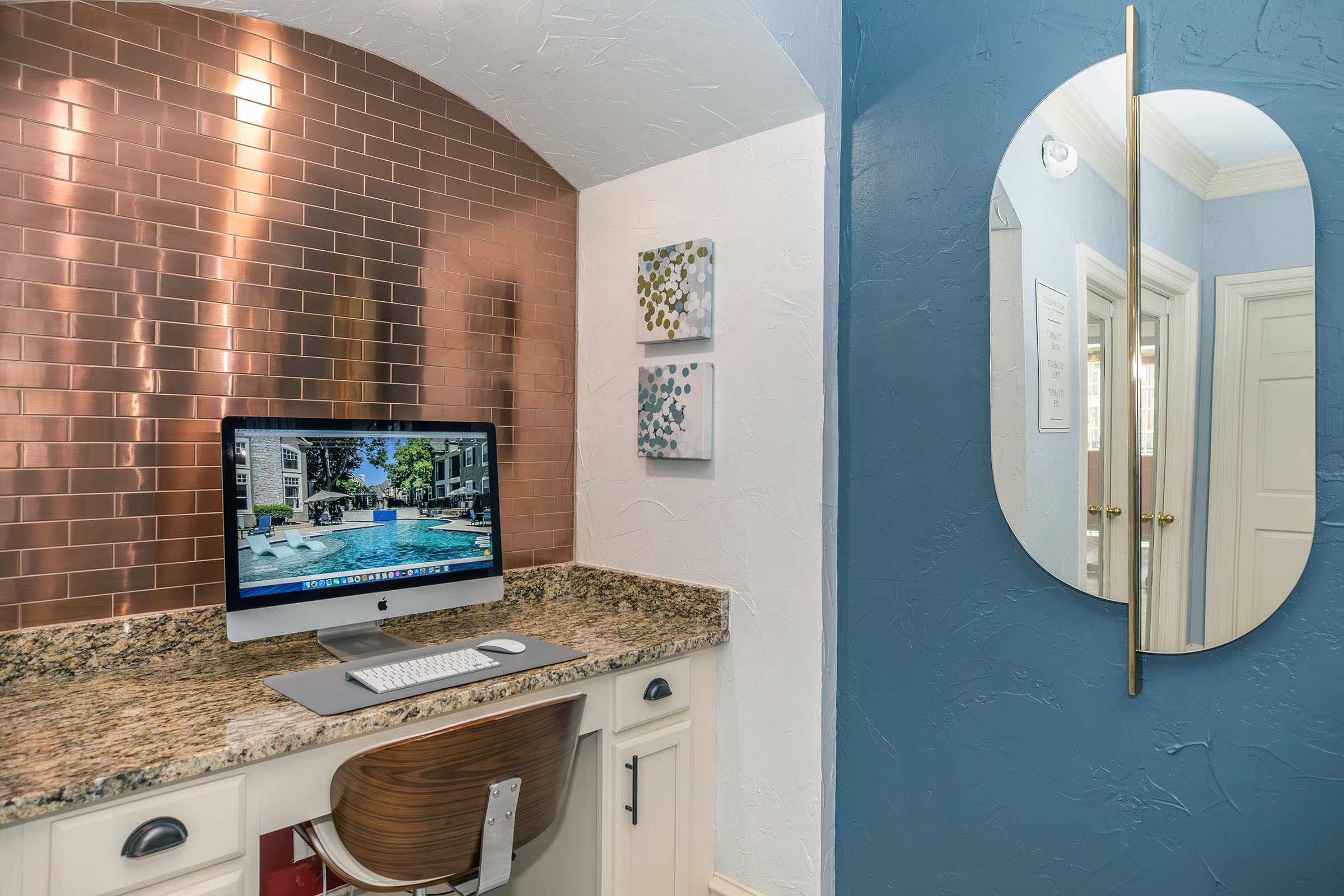 A stylish workspace featuring a modern computer on a granite countertop, with a copper backsplash. The wall is painted blue, and a large, oval mirror reflects light. On the wall, there are two small abstract artworks. The overall aesthetic is contemporary and inviting.