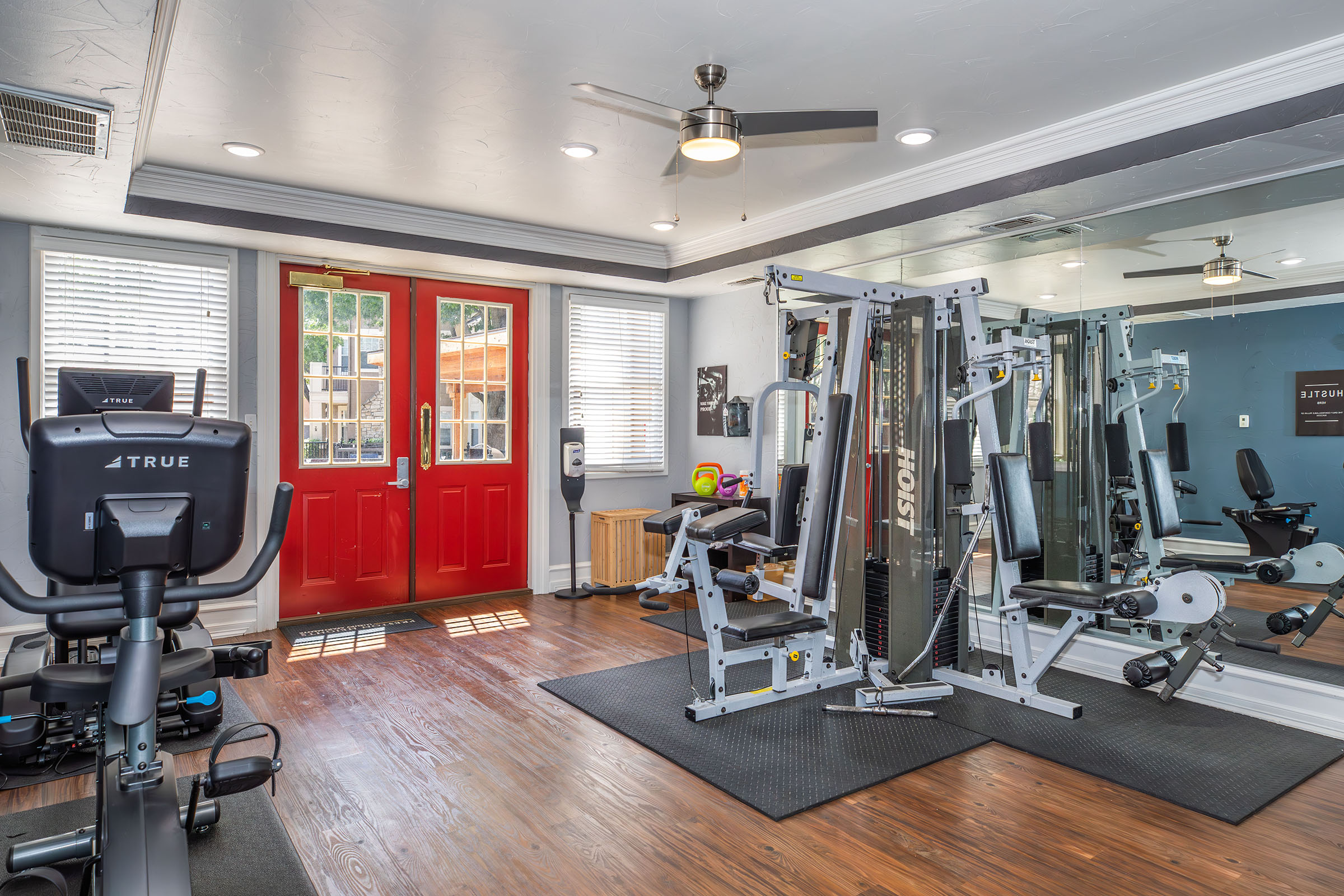 Interior view of a modern gym featuring exercise equipment like a stationary bike, weight machines, and a mirror. The room has wooden floors, blue walls, and abundant natural light from windows. Two red entry doors create a welcoming atmosphere. A fan is mounted on the ceiling for ventilation.