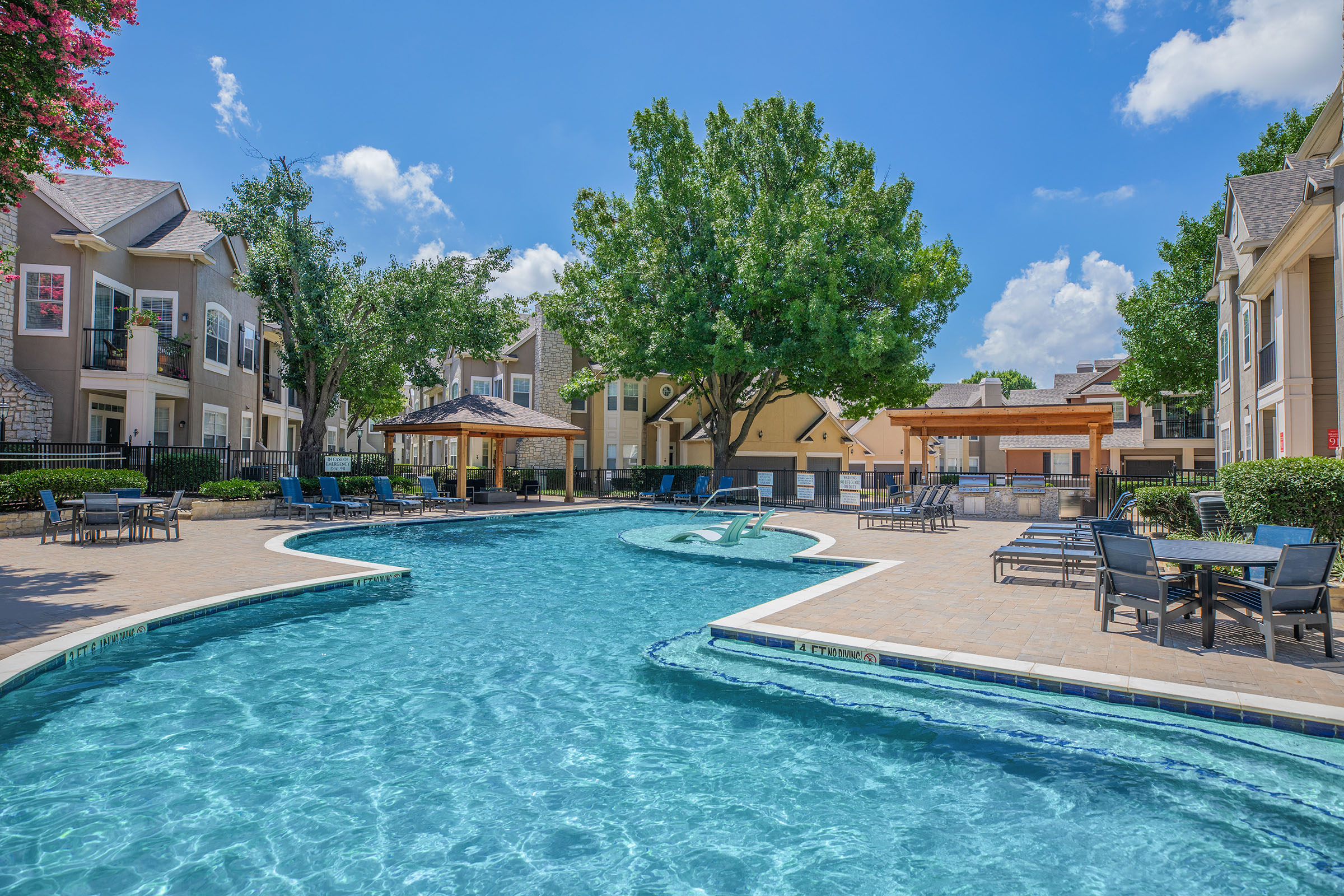 A spacious swimming pool surrounded by lounge chairs and shaded areas, set within a residential complex. Lush trees and bright blue skies enhance the inviting atmosphere. The pool features a curvy design with a shallow area, making it ideal for relaxation and recreation.