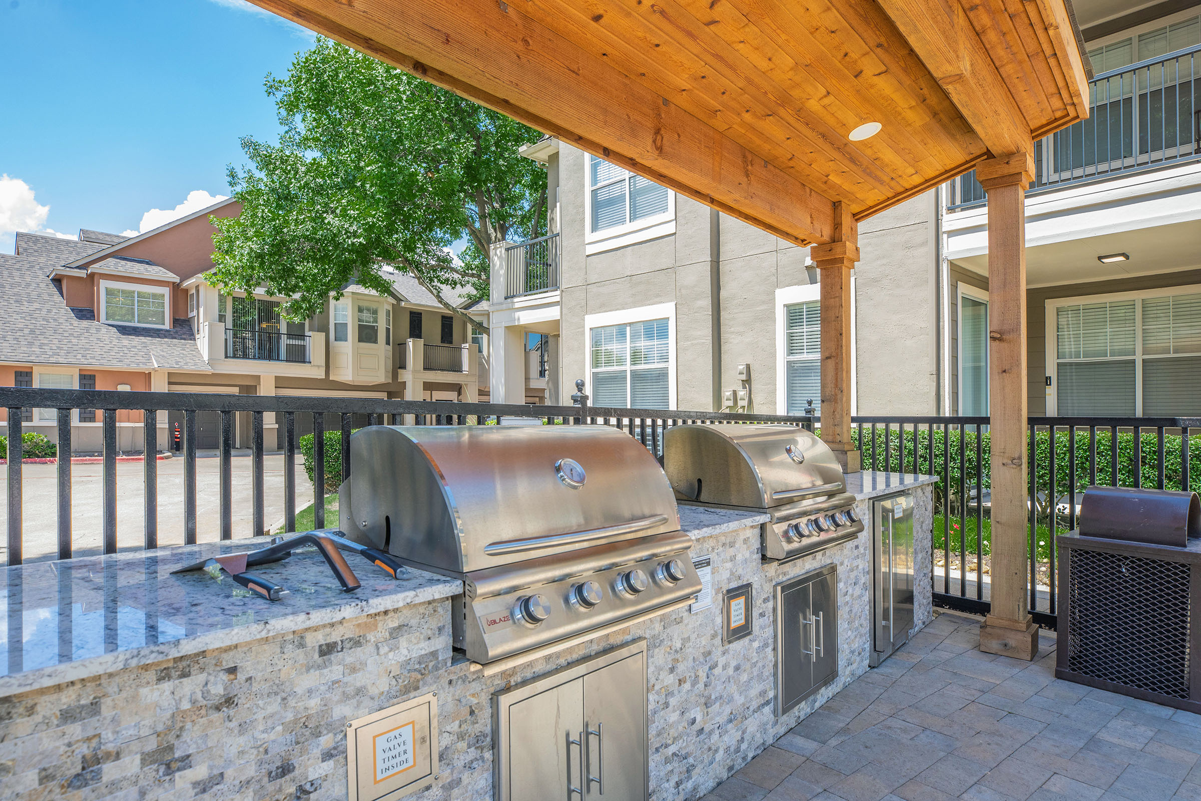 Outdoor grilling area featuring three stainless steel gas grills, granite countertops, and a wooden pergola overhead. The scene is set in a spacious courtyard of an apartment complex, with well-maintained greenery and modern buildings in the background under a clear blue sky.