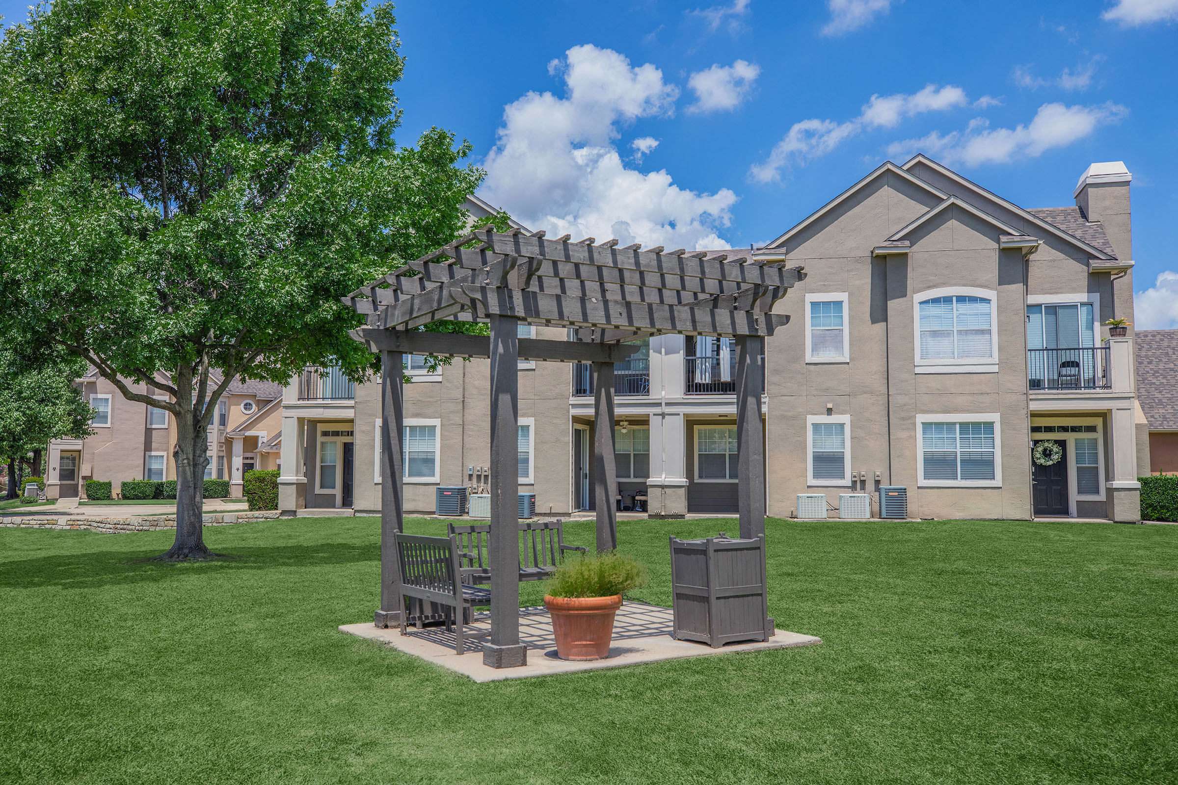 A landscaped area featuring a wooden pergola with benches and potted plants, surrounded by green grass. In the background, there are two multi-story apartment buildings with windows and doors. The sky is partly cloudy with blue and white clouds.