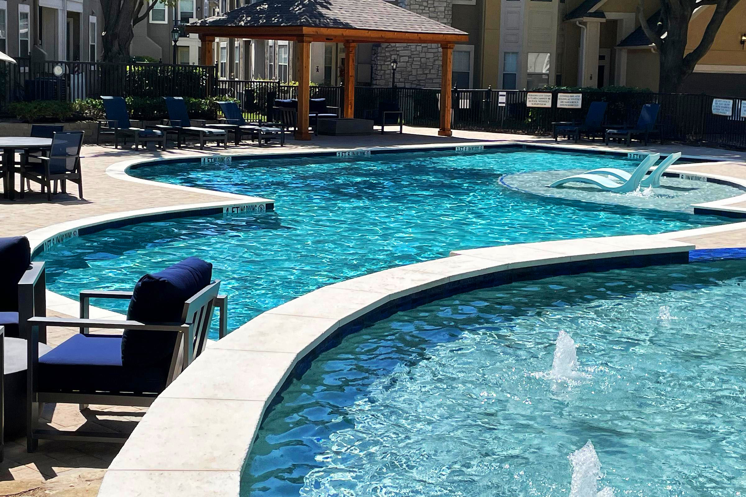 A sunny pool area featuring a curved swimming pool with clear blue water, surrounded by lounge chairs and tables. In the background, there is a gazebo and a landscaped area. Fountains are visible in the pool, adding to the relaxing atmosphere. Great for leisure and relaxation.