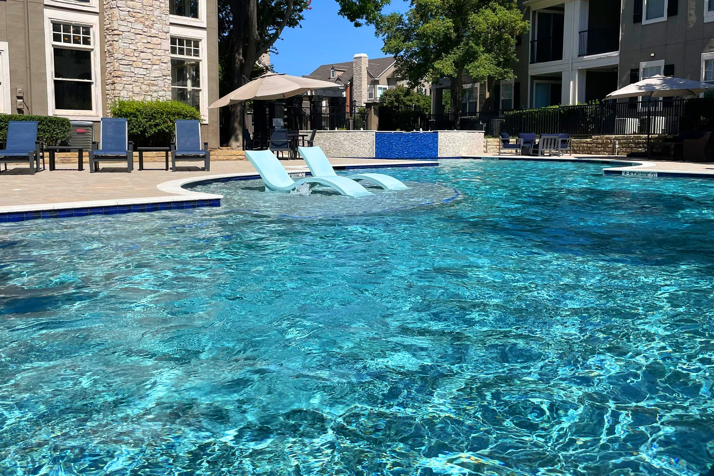 A serene pool scene featuring clear, blue water with two floating lounge chairs partially submerged in the water. Surrounding the pool are sun loungers and umbrellas, with a backdrop of modern apartment buildings and lush green trees under a bright blue sky.