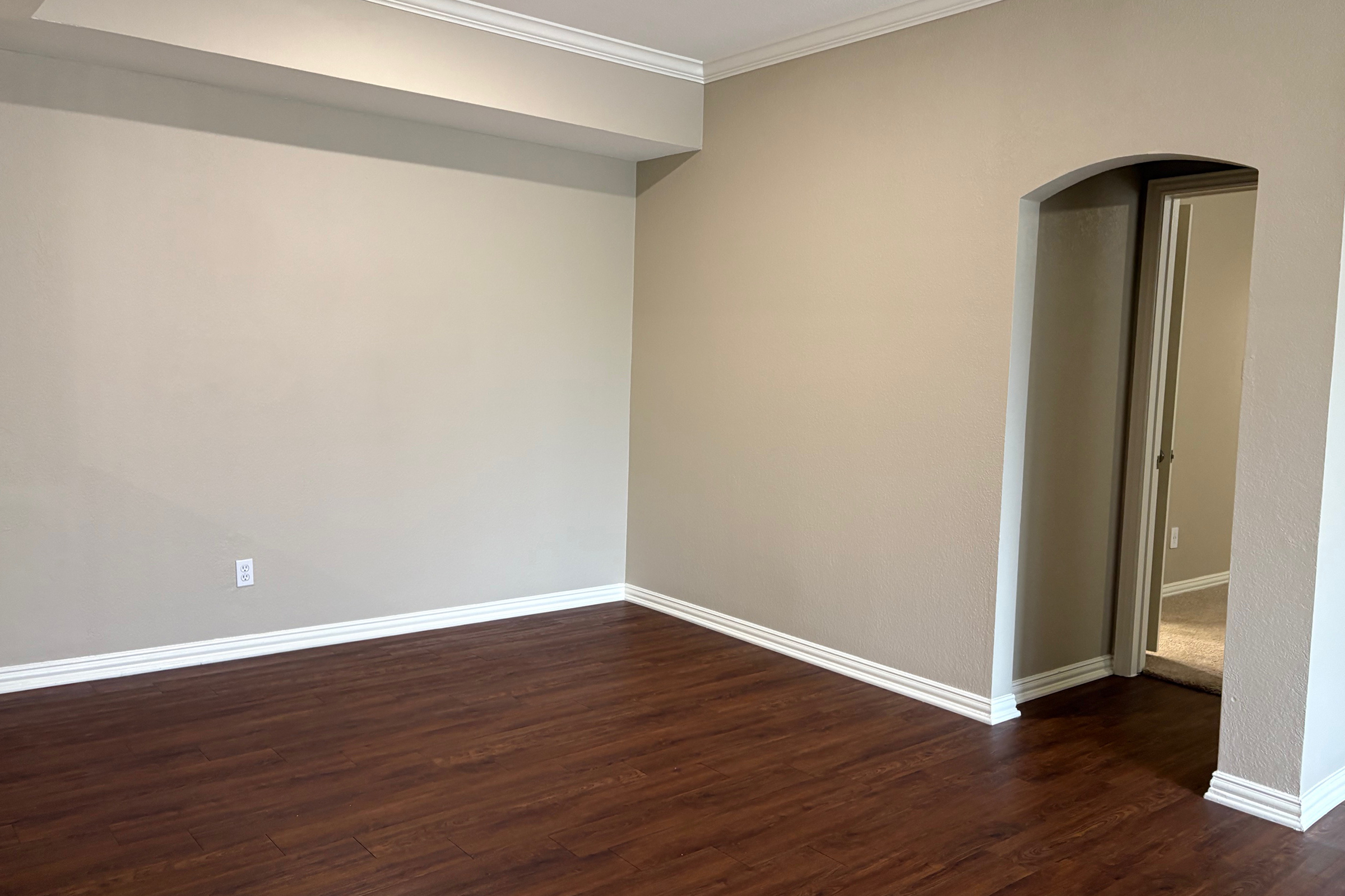 Empty room featuring light brown walls and a hardwood floor. It has a small arch leading to a doorway on the right, and no furniture is present. Natural light may be coming through the doorway, highlighting the simplicity of the space.