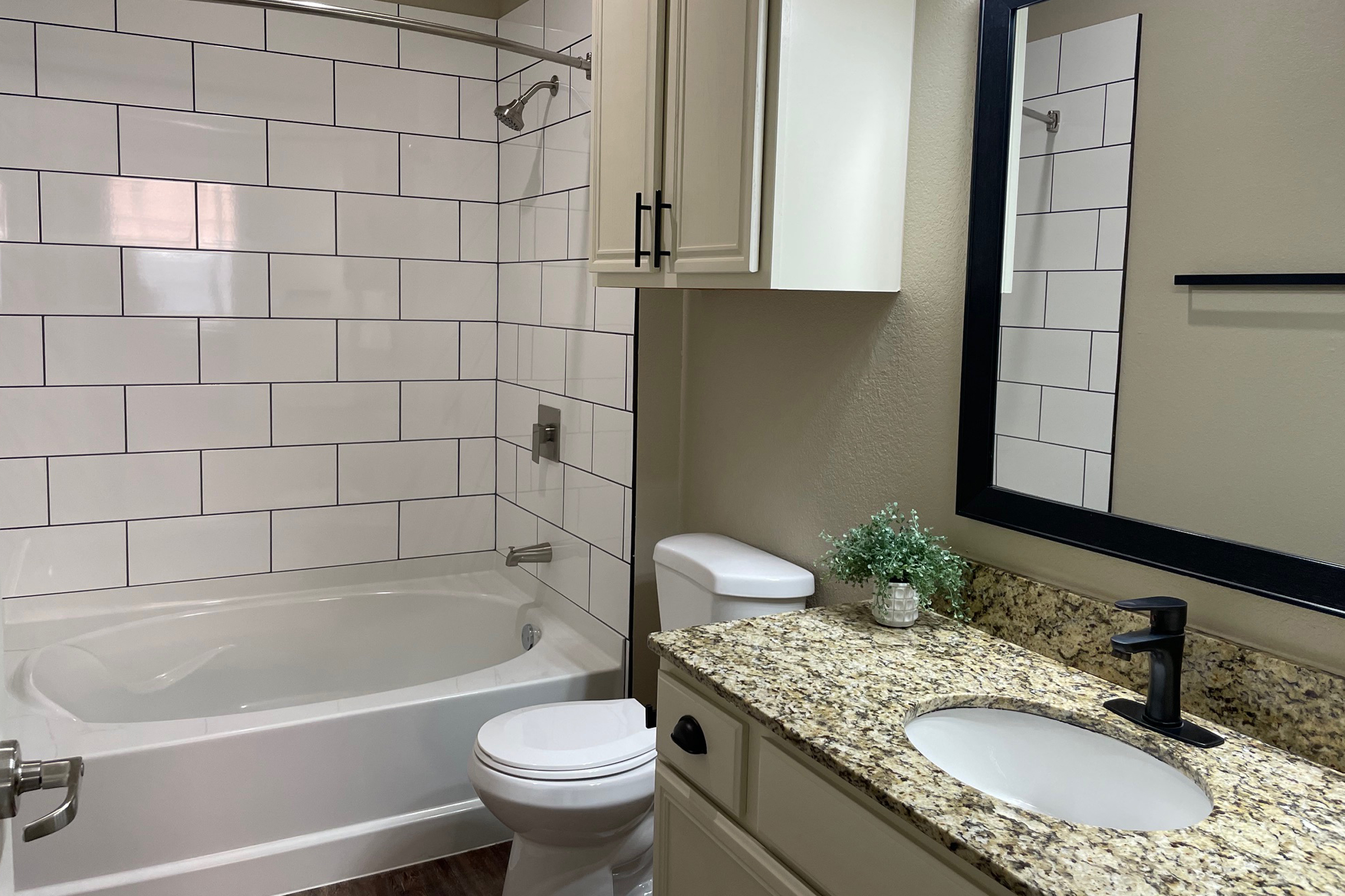 A modern bathroom featuring a bathtub with a shower, a white toilet, and a granite countertop with a sink. The walls have white tiles arranged in a grid pattern, and there is a small potted plant on the countertop. A large mirror is mounted above the sink, reflecting the room's neutral tones.